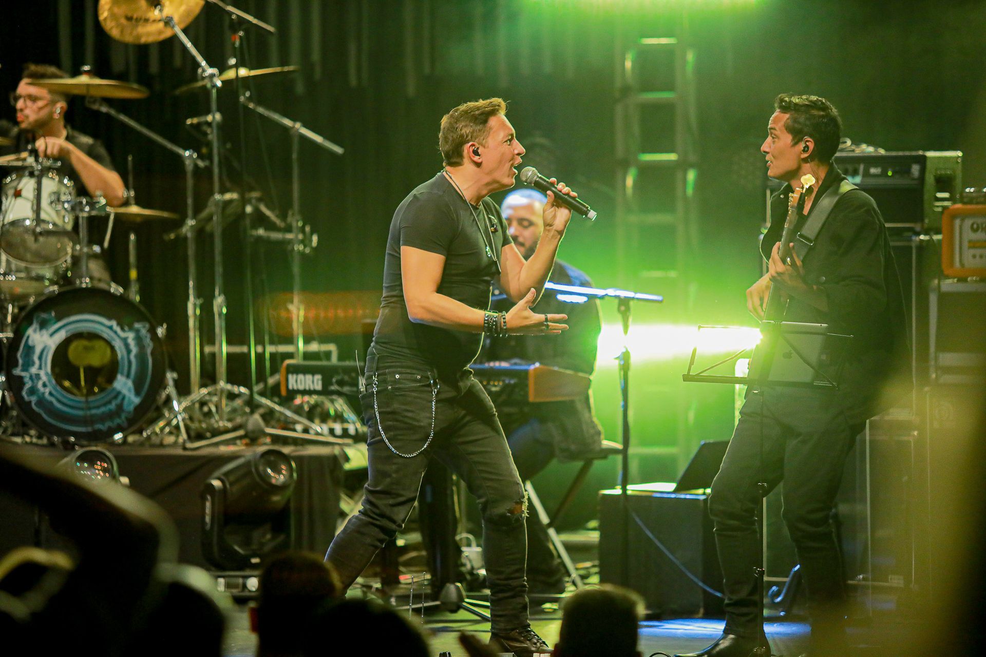Uma banda se apresenta no palco sob luzes verdes, com o vocalista usando um microfone está de frente para um guitarrista.