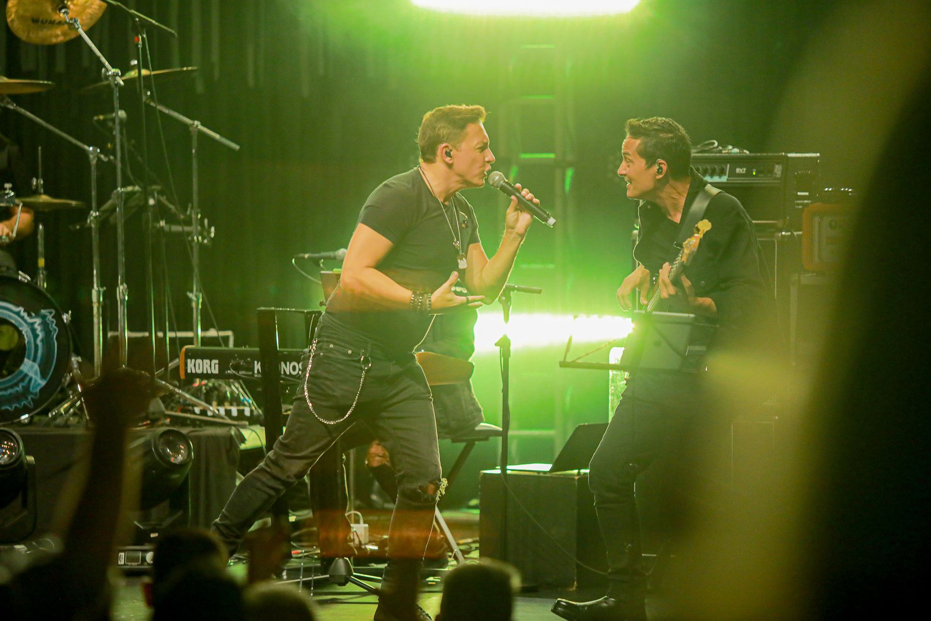 Dois músicos se apresentam em um palco iluminado por uma luz verde brilhante; um canta enquanto o outro toca guitarra.