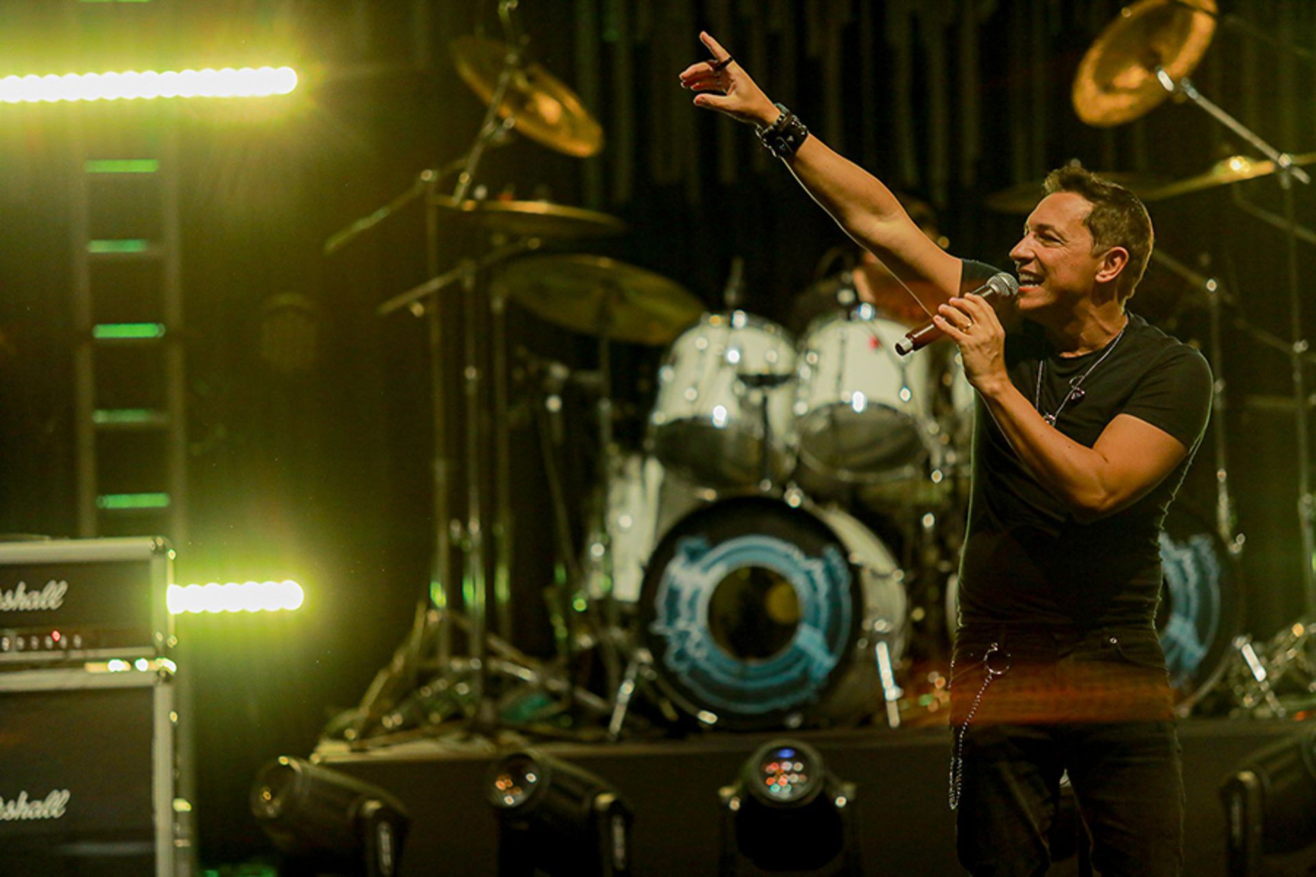 Um artista cantando em um microfone no palco, apontando um dos braços para cima, com uma bateria ao fundo.