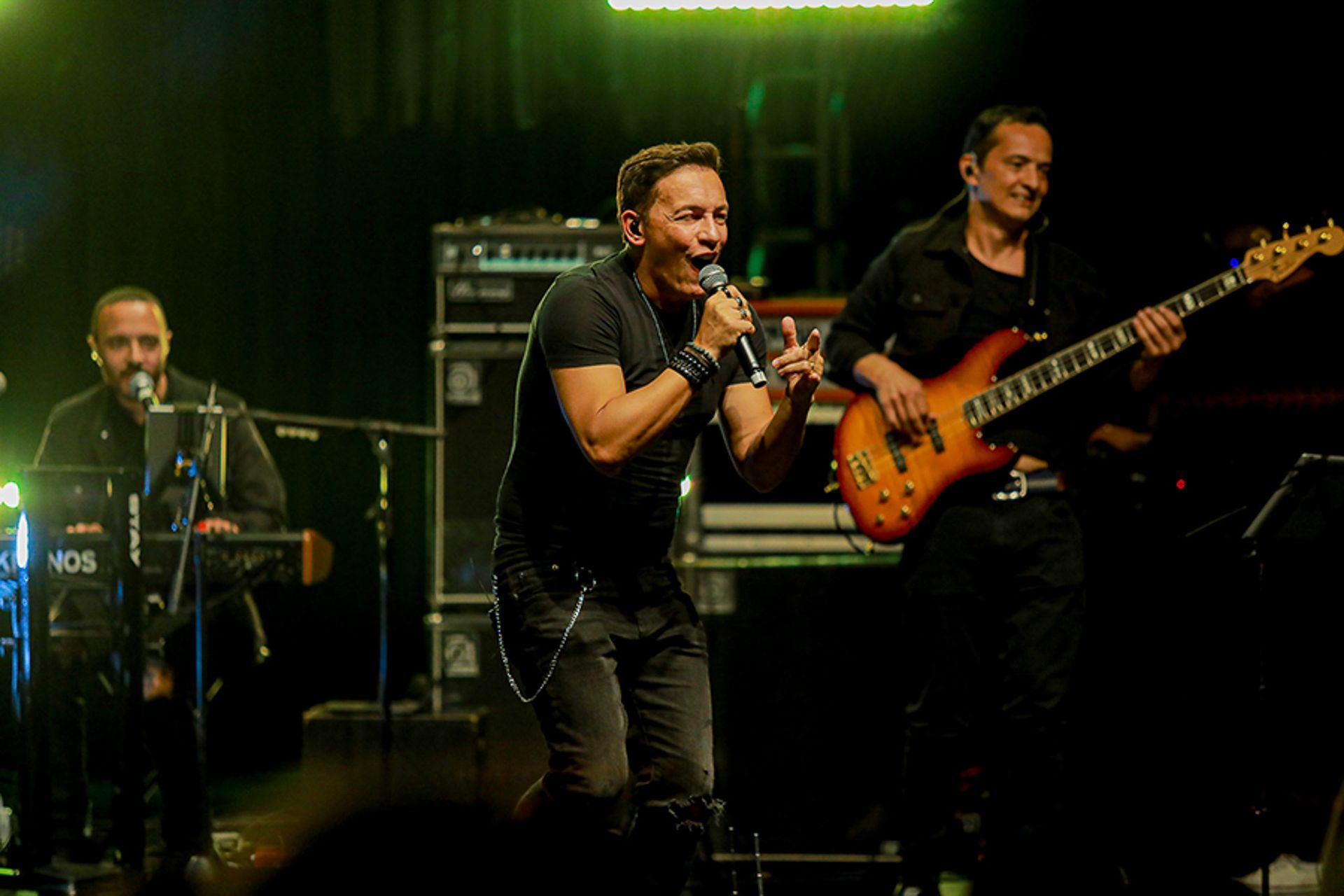 Uma banda se apresenta no palco: um vocalista canta ao microfone acompanhado por um tecladista e um baixista.