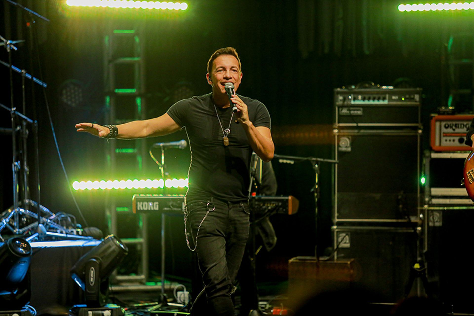 Um artista vestindo camisa preta e calça jeans segura um microfone e gesticula em um palco de concerto iluminado.