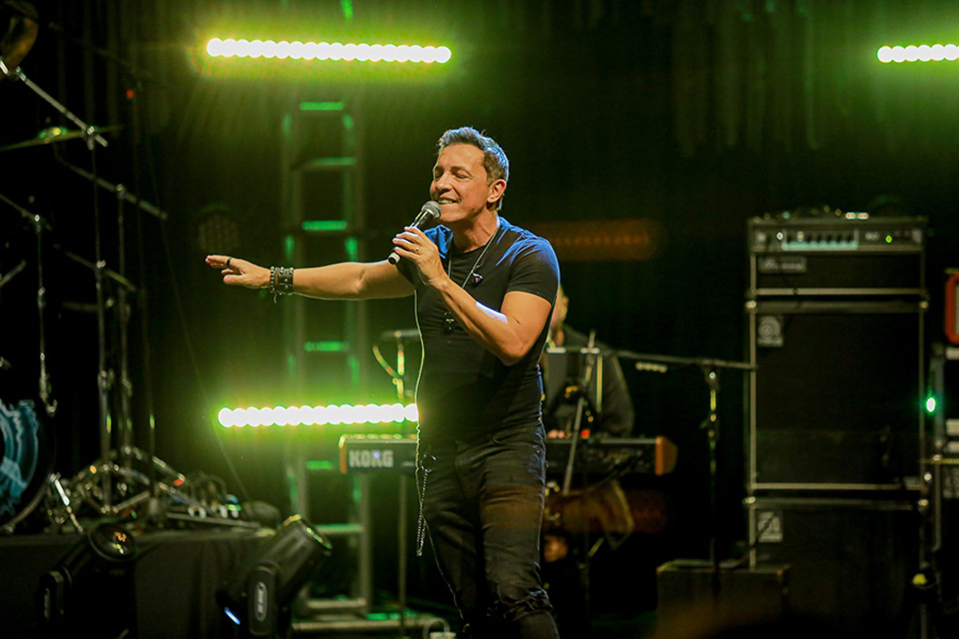 Um artista canta em um microfone em um palco com iluminação verde e equipamentos musicais ao fundo.