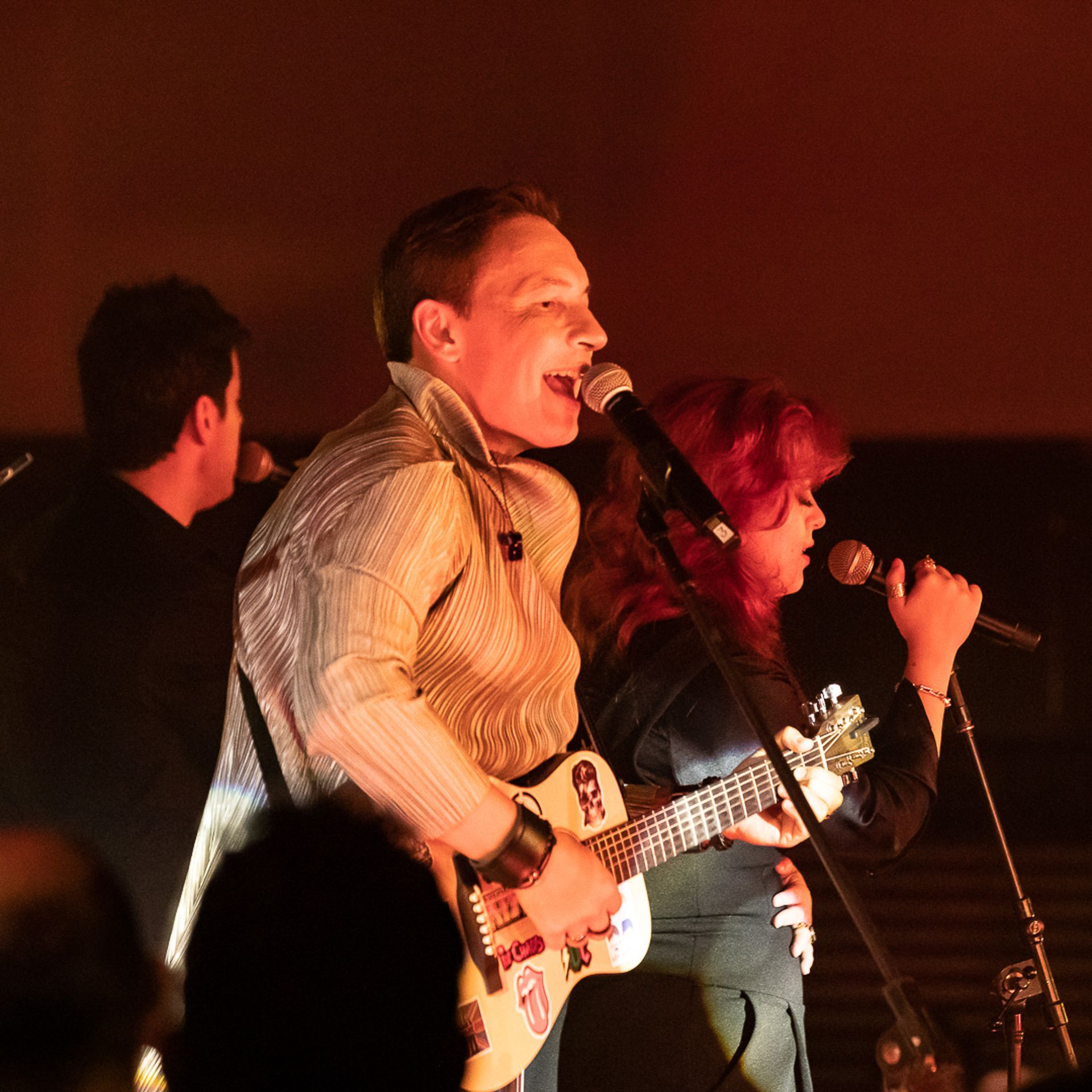 Um músico com uma camisa estampada toca guitarra e canta ao microfone ao lado de uma vocalista em um palco escuro.