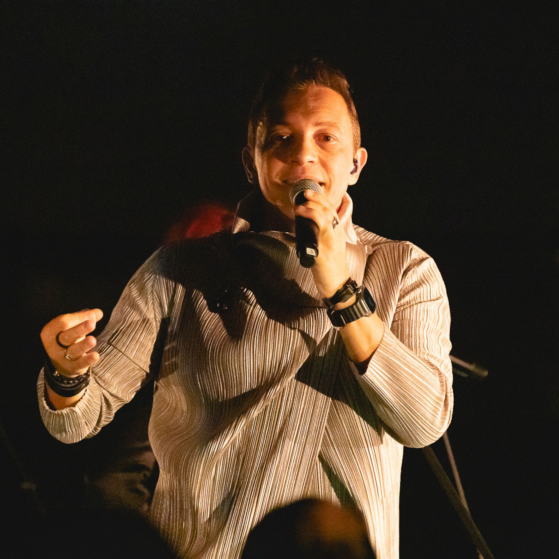 Um artista vestindo uma camisa de mangas compridas estampada, segurando um microfone e cantando em um palco escuro.