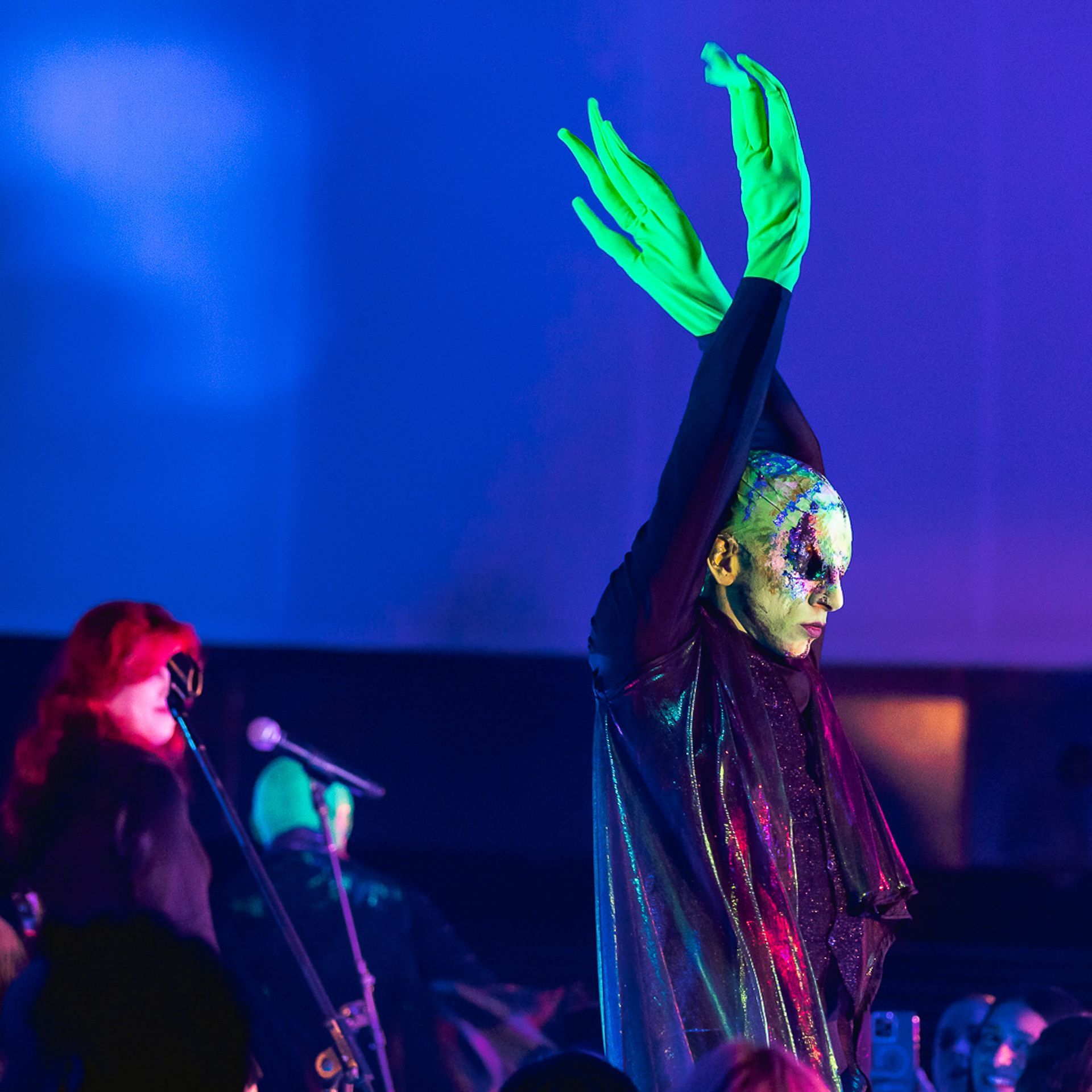 Um artista com maquiagem facial estampada e luvas verde-brilhantes erguidas acima da cabeça em um palco com iluminação azul.