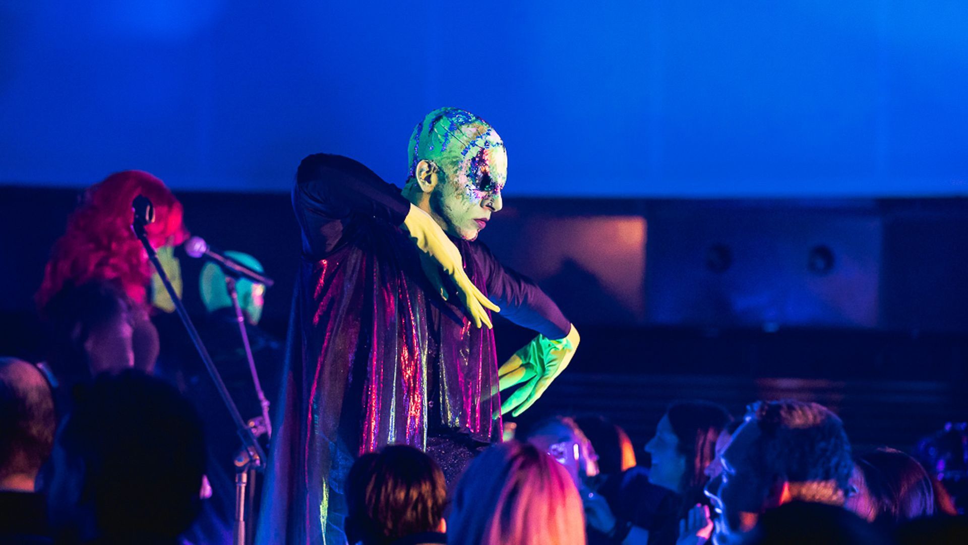 Um artista com o rosto pintado e um adorno de cabeça estampado usa uma capa escura e luvas coloridas no palco