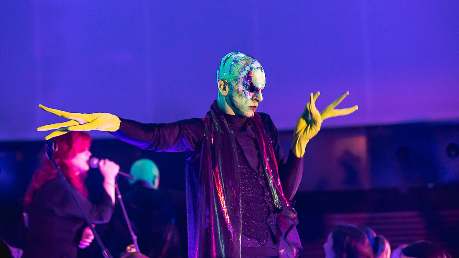 Um artista com maquiagem estilizada e luvas amarelas posa em um palco sob iluminação roxa, com uma banda ao fundo.