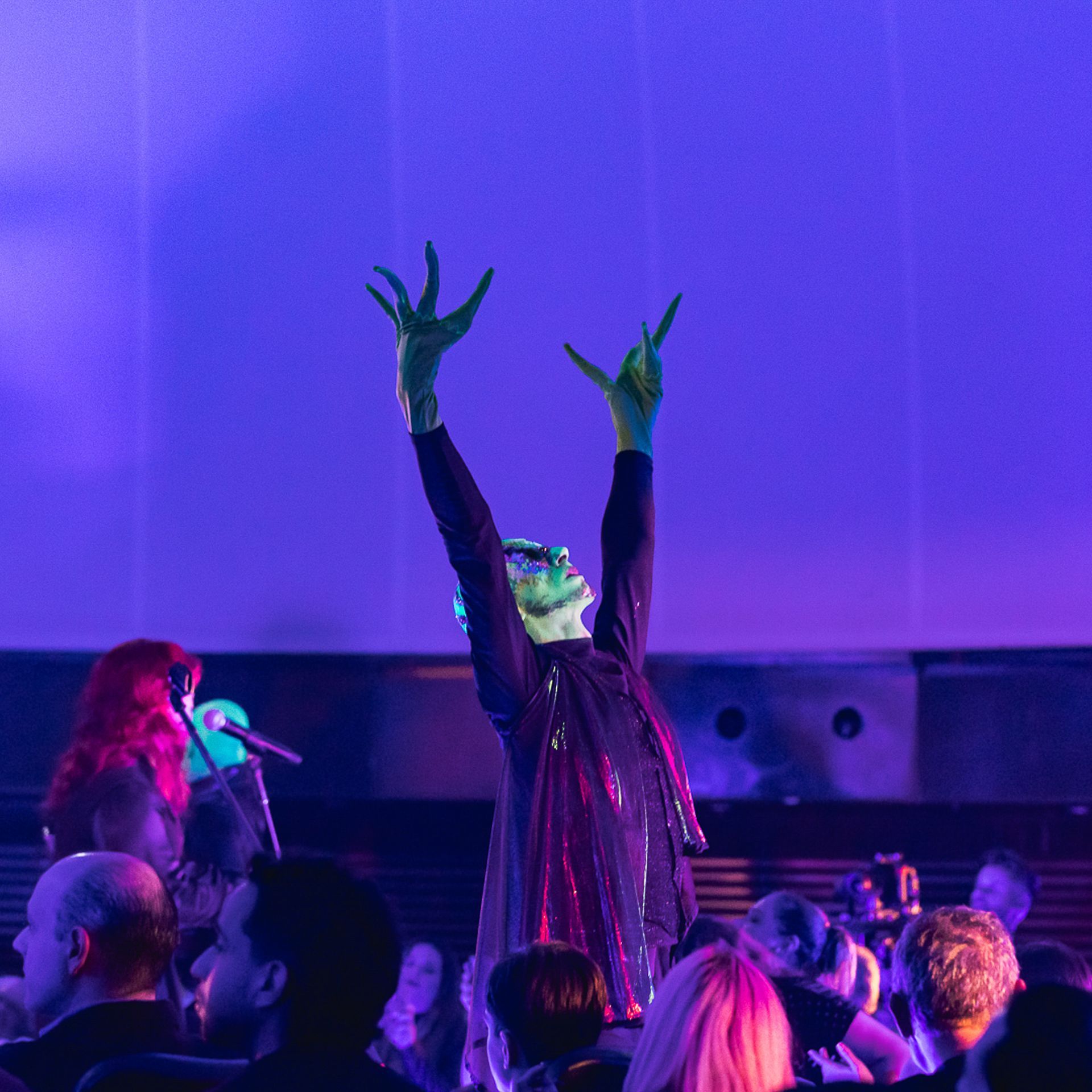 Um artista com a pele esverdeada e as mãos erguidas está em um palco banhado por uma luz roxa intensa.