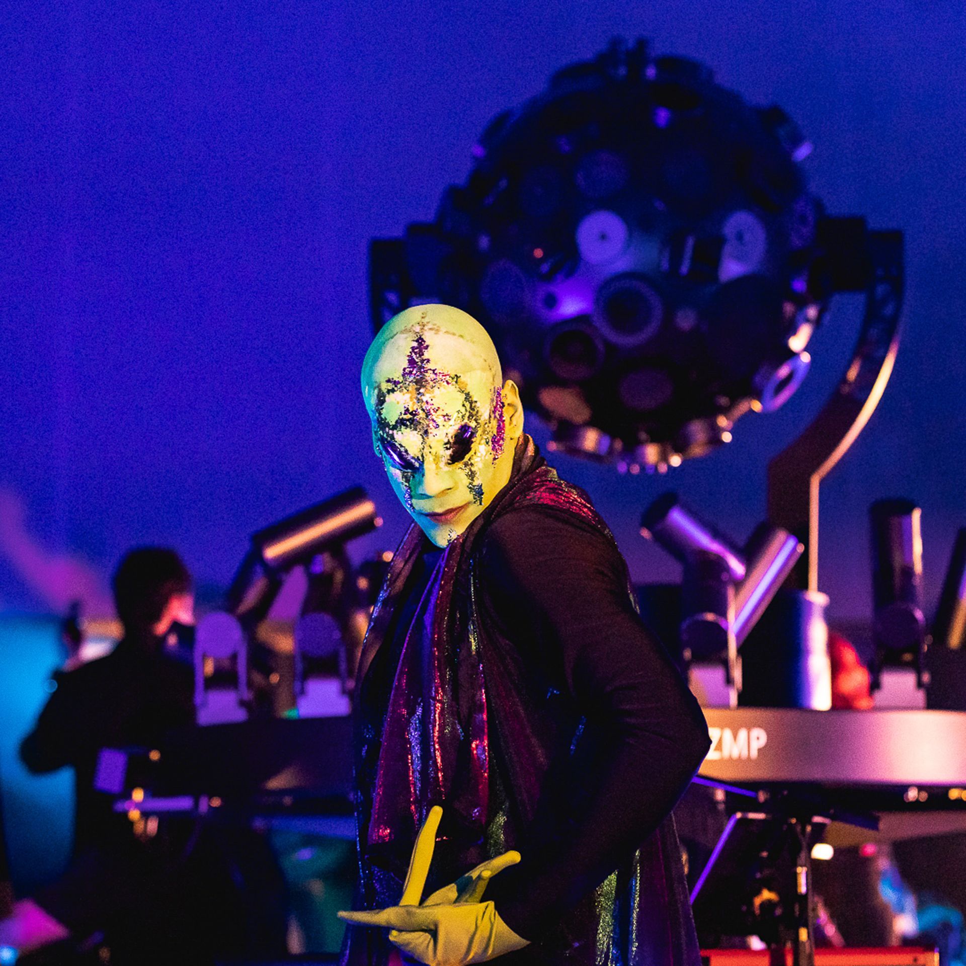 Um artista com uma máscara facial pálida e decorada e luvas verdes posa no palco vestida de escuro contra uma luz azul.
