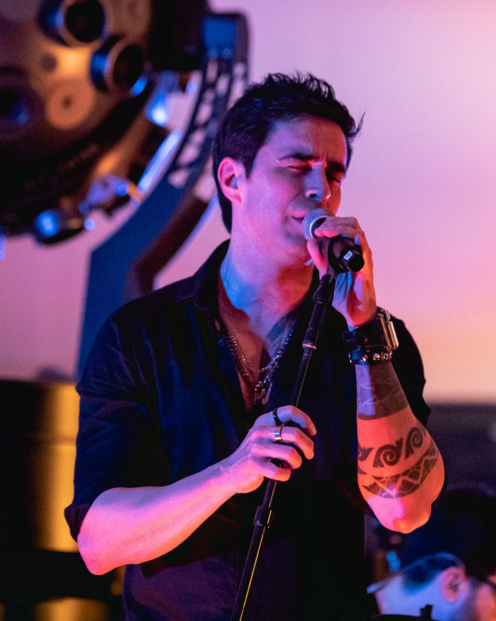 Um artista com tatuagens no braço cantando em um microfone no palco sob iluminação rosa e azul.