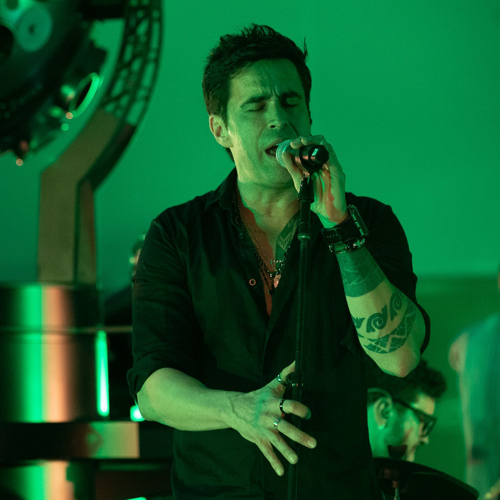 Um artista de camisa preta cantando em um microfone no palco, banhado por luz verde, com uma tatuagem visível no braço.