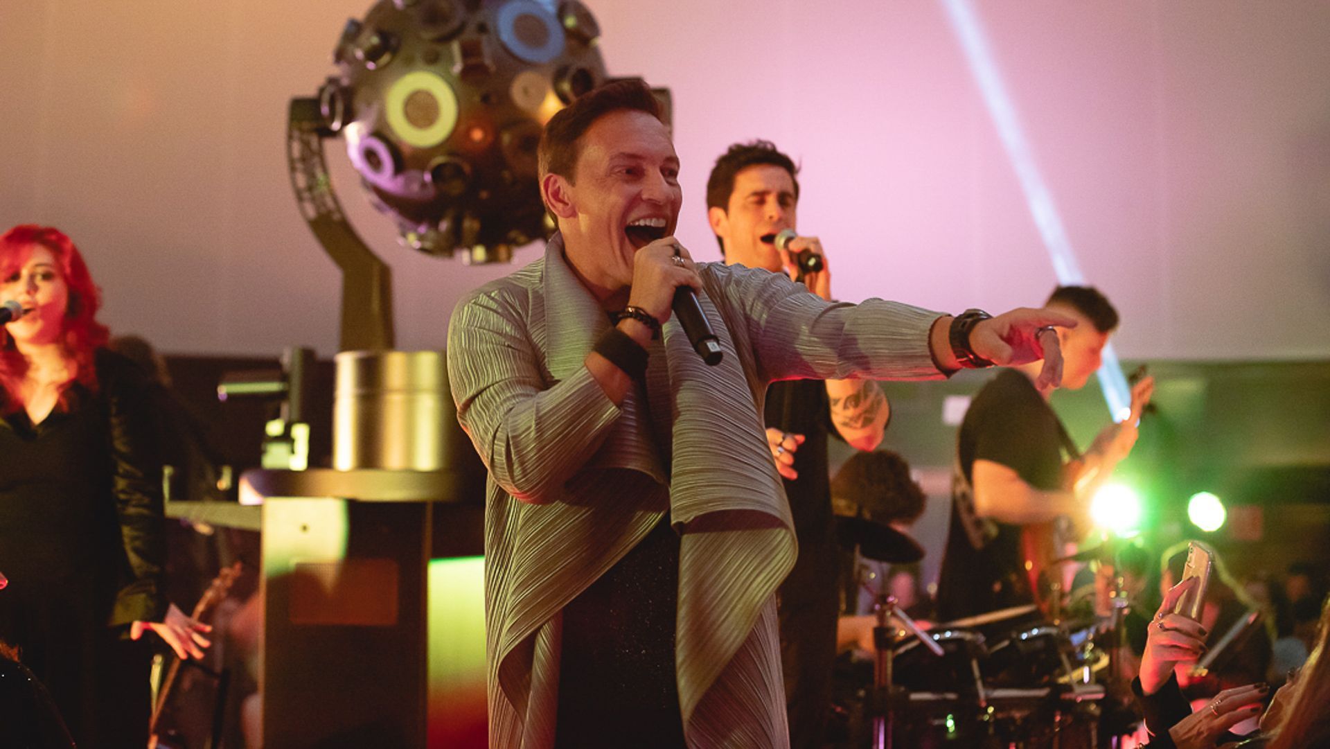 Uma banda se apresenta no palco com um vocalista gesticulando para a plateia, iluminada por luzes de palco coloridas.
