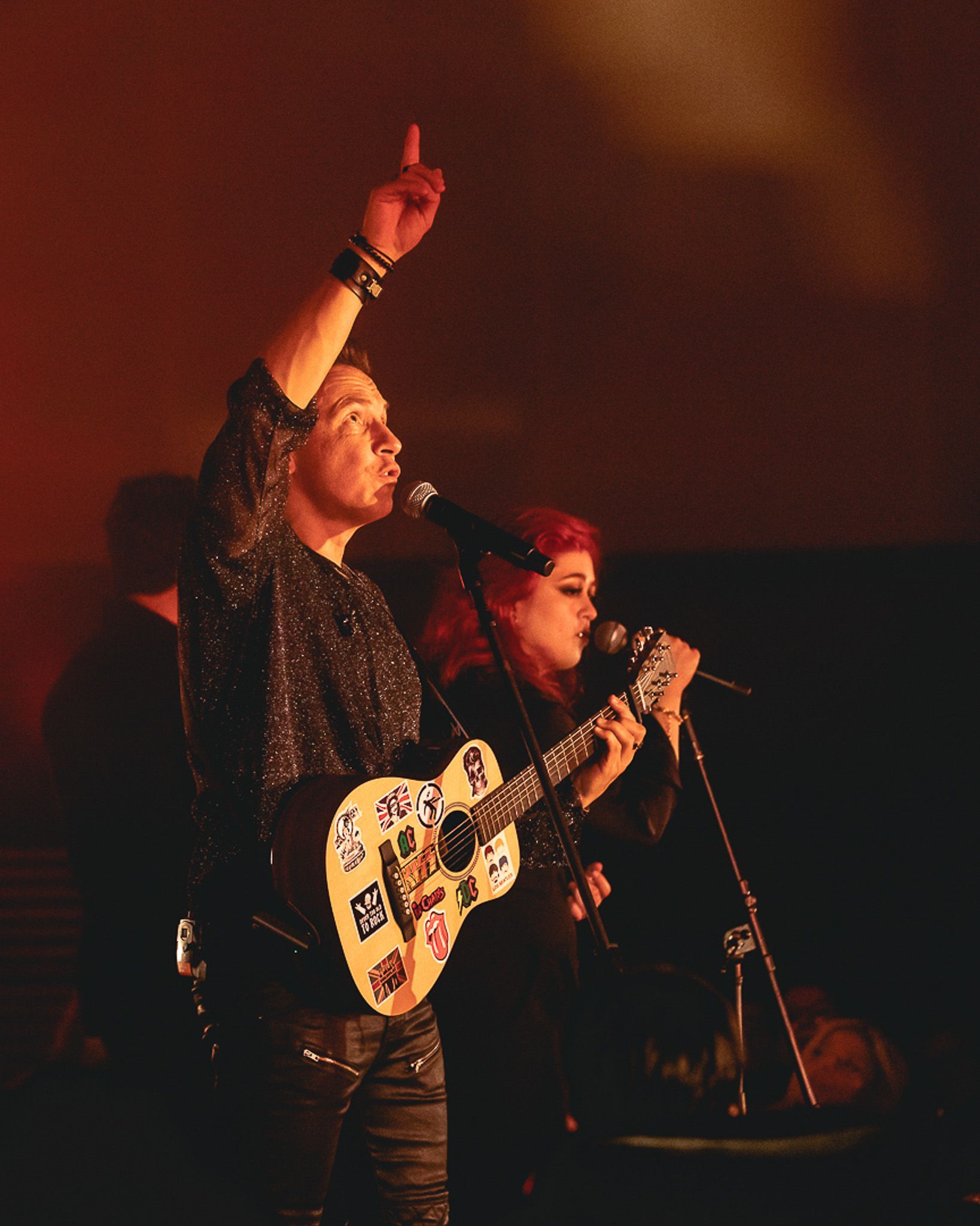 Um músico tocando um violão coberto de adesivos e gesticulando para cima enquanto canta com uma backing vocal no palco.
