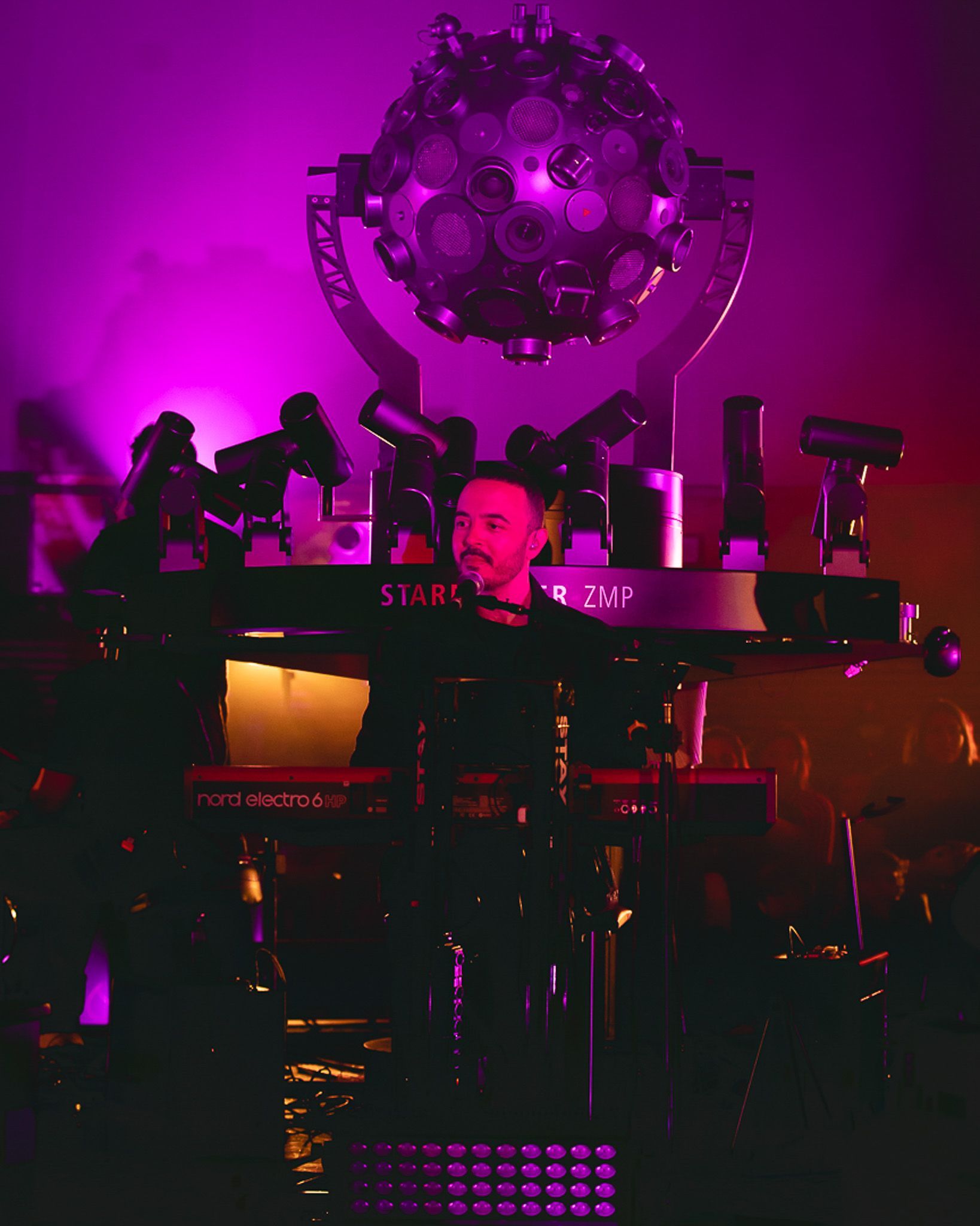 Um artista tocando teclado em um palco de concerto, iluminado por luz roxa com um grande refletor esférico acima dela.