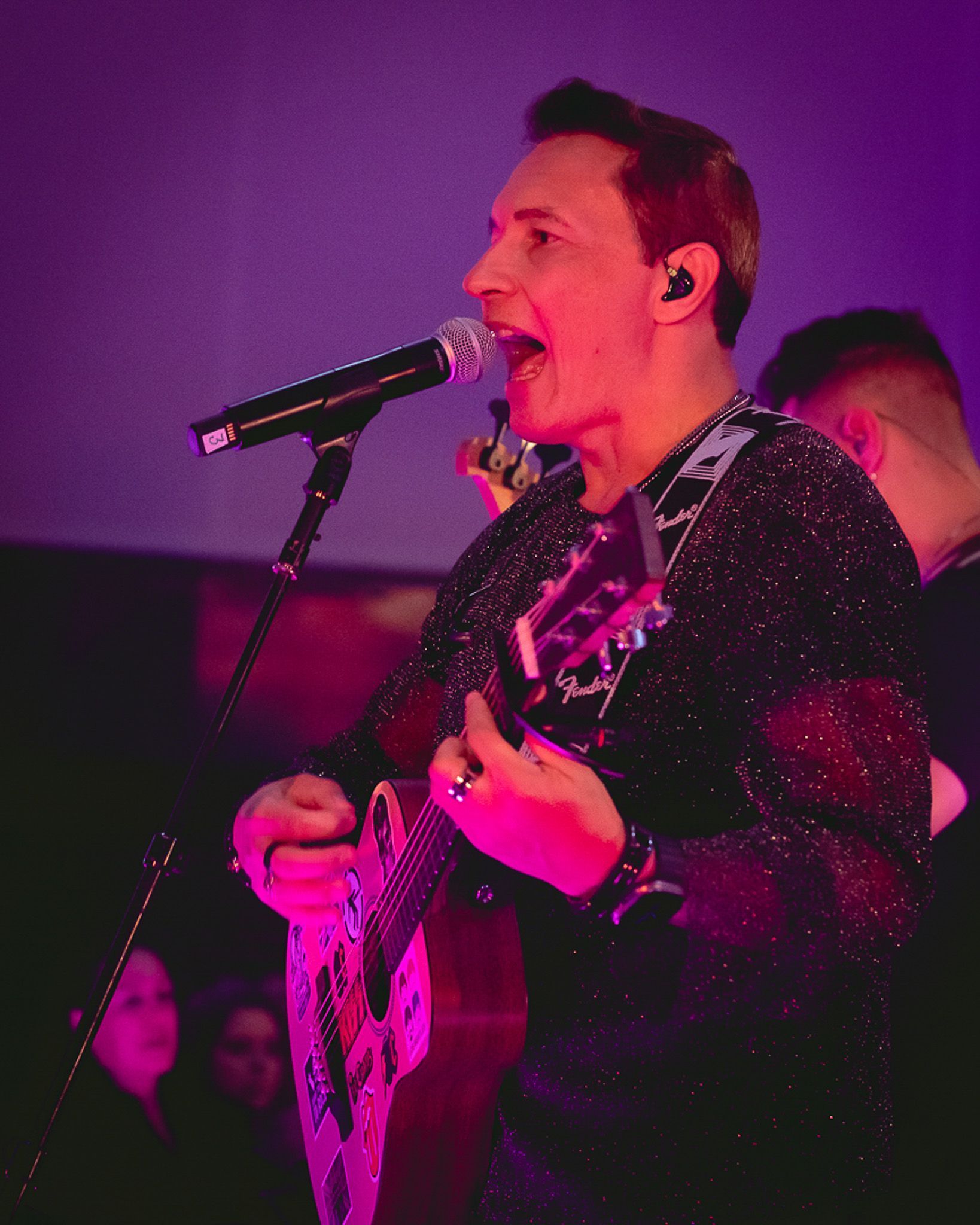 Um artista cantando ao microfone enquanto toca violão, iluminada por luzes de palco roxas.