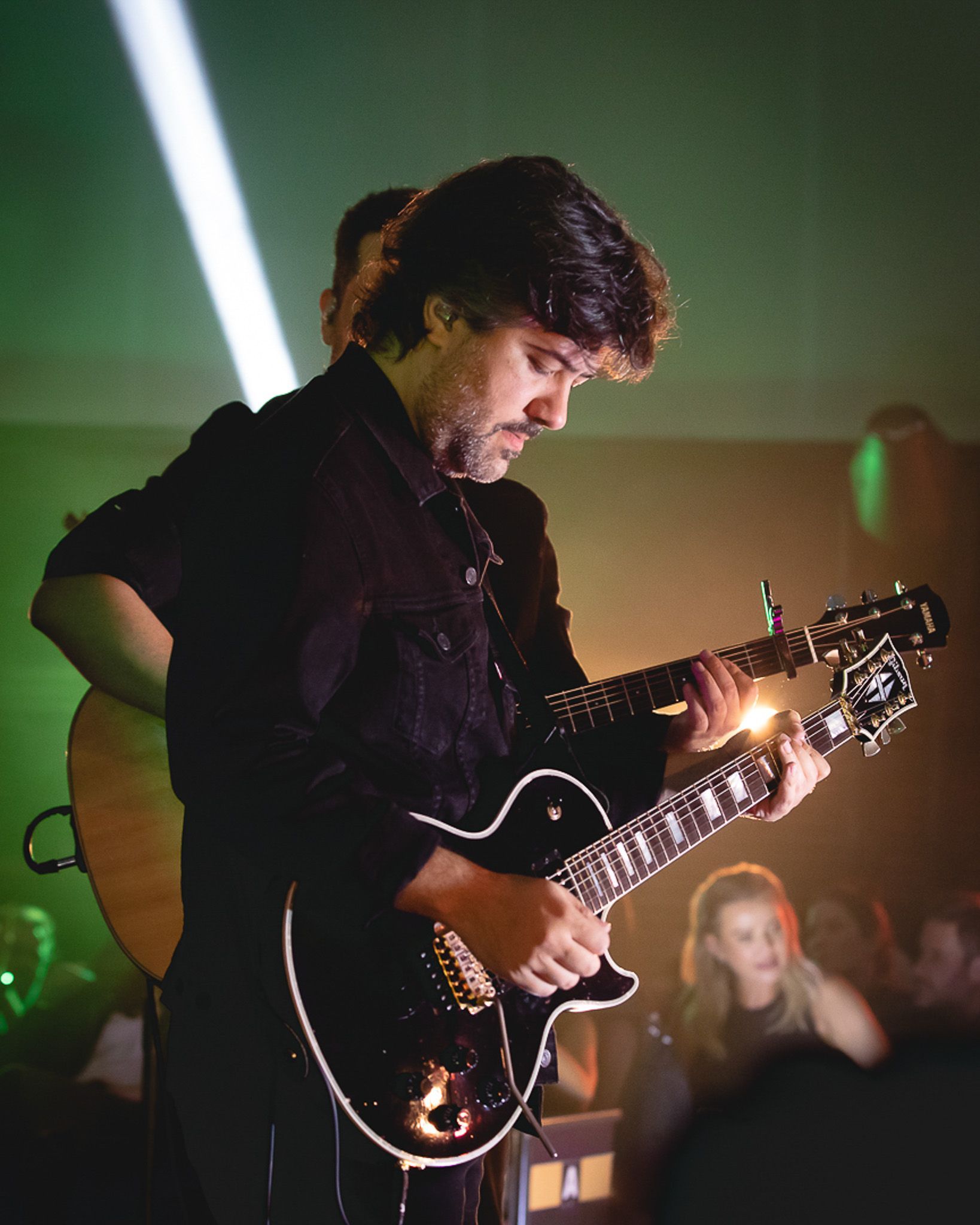 Um artista toca guitarra elétrica intensamente no palco, com uma plateia desfocada visível ao fundo.