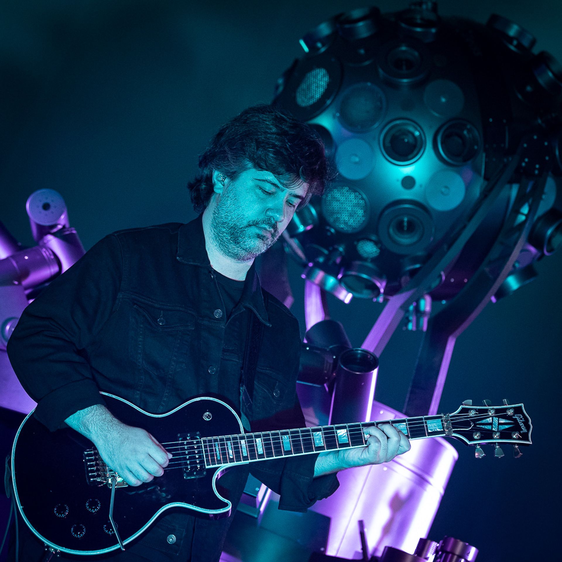 Um músico toca guitarra no palco sob uma iluminação azul fria, posicionado em frente a uma grande escultura esférica.