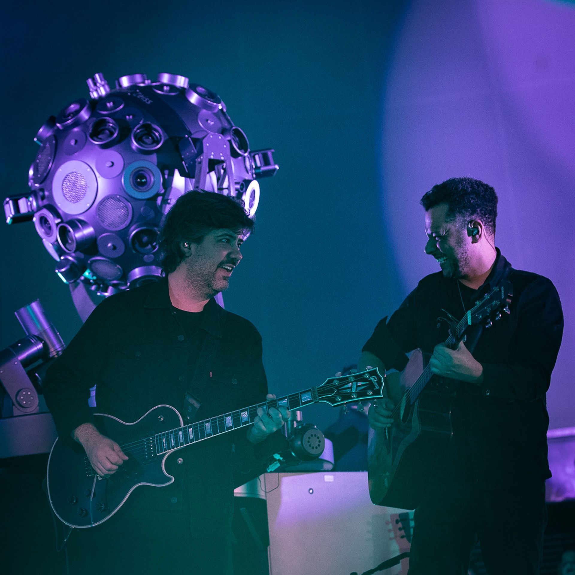Dois músicos se apresentam no palco com guitarras em frente a um grande refletor esférico, banhado por luz roxa.