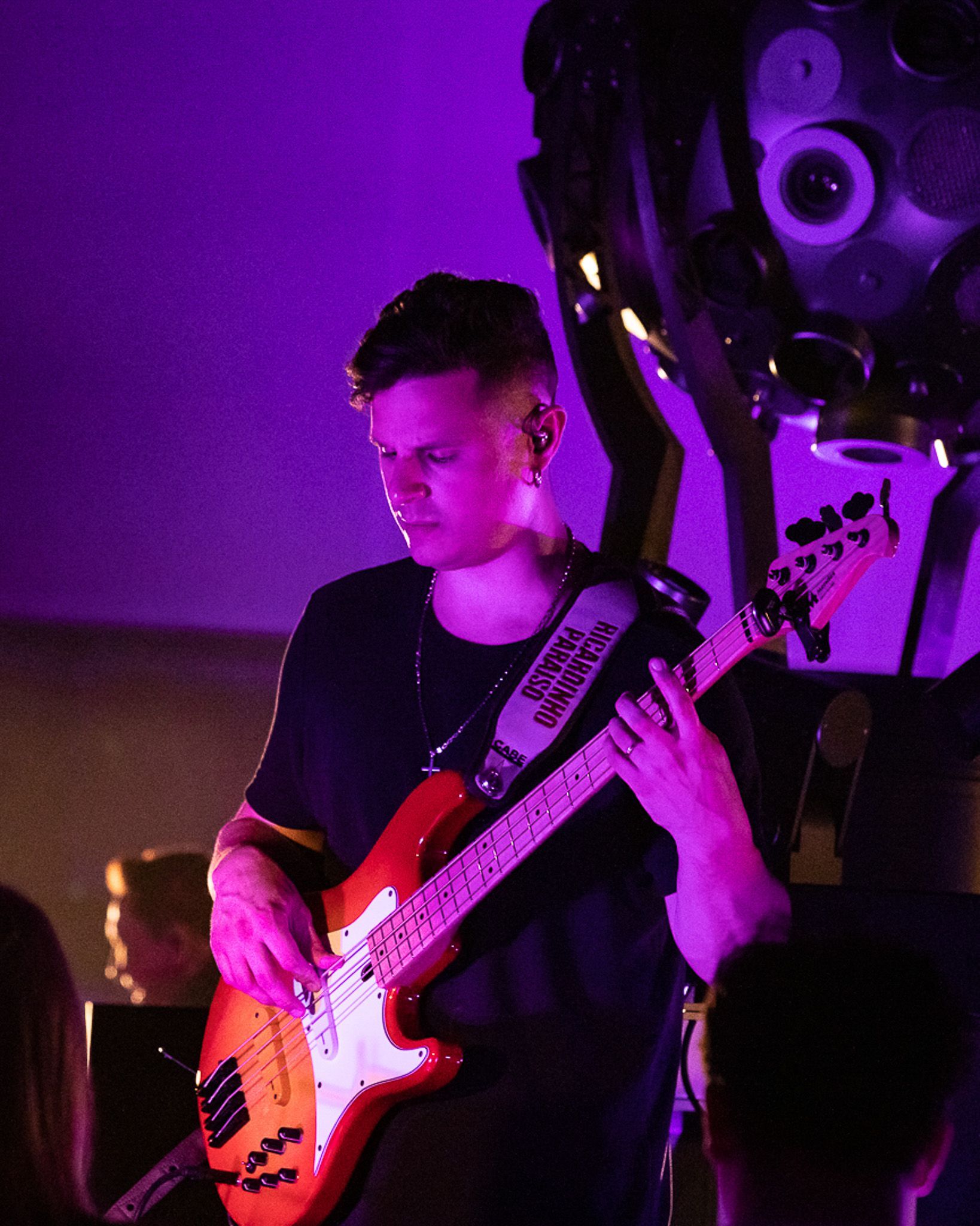 Um músico toca um baixo elétrico vermelho sob uma vibrante iluminação de palco roxa, com uma luz de palco visível ao fundo.