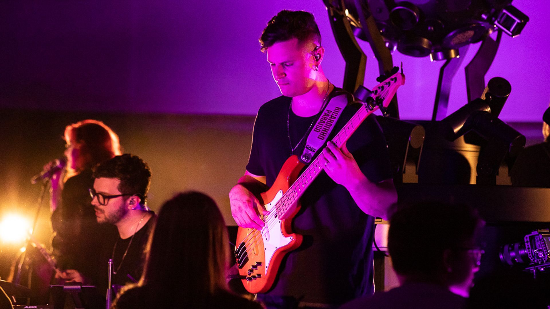 Uma banda se apresentando em um palco sob iluminação roxa, com um baixista tocando uma guitarra em primeiro plano.