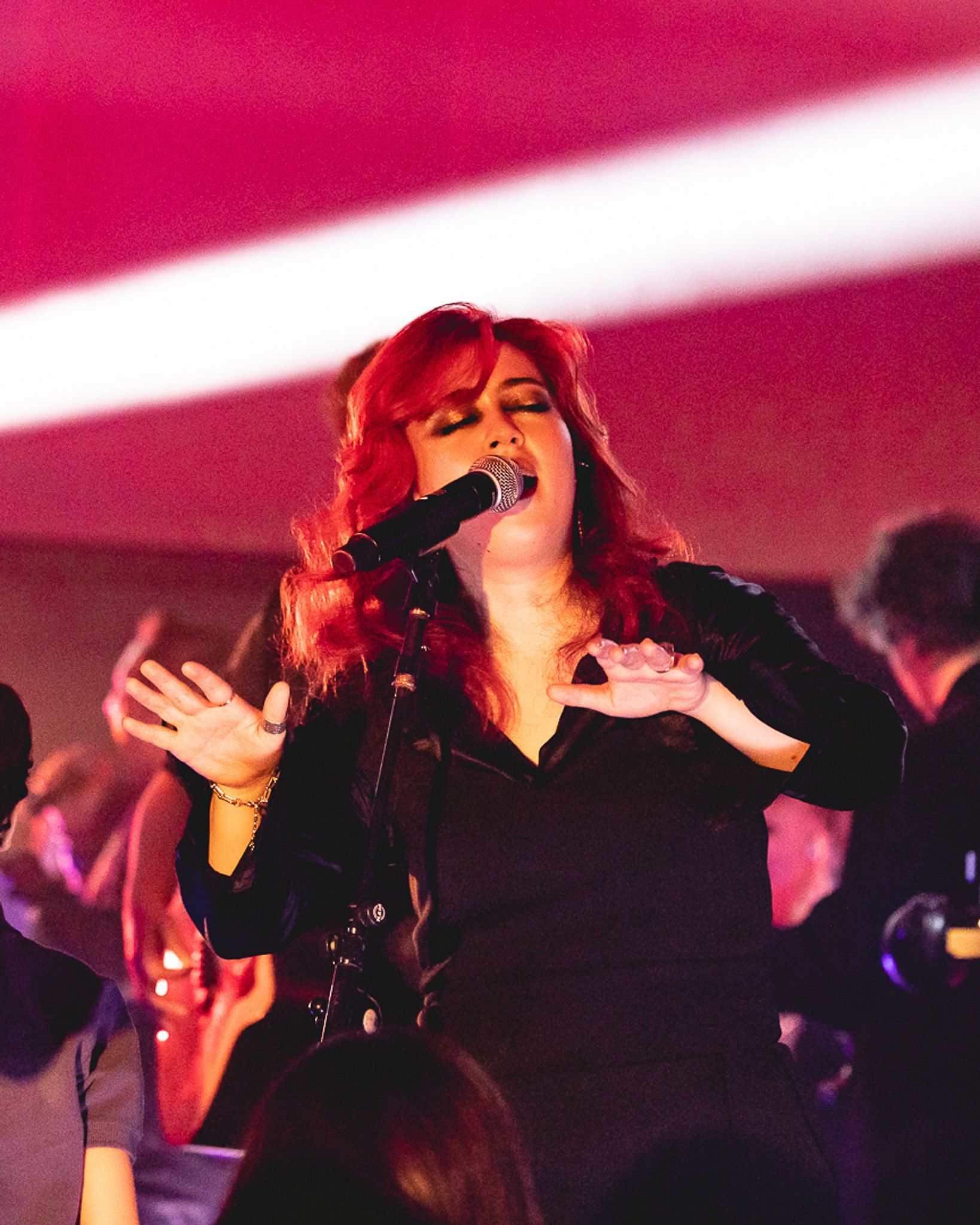 Uma artista com cabelo vermelho vivo cantando em um microfone no palco, gesticulando com as mãos sob uma iluminação rosa.