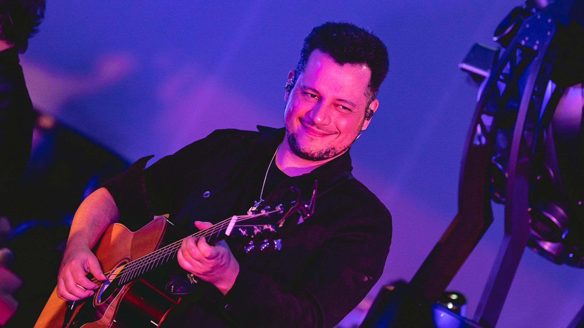 Um músico sorridente tocando violão em um palco iluminado por uma vibrante luz roxa.