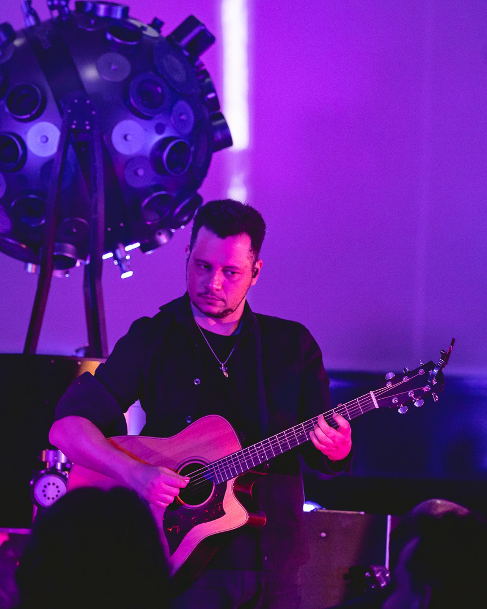 Um músico tocando violão no palco sob iluminação roxa, em frente a uma grande estrutura de luzes circular.