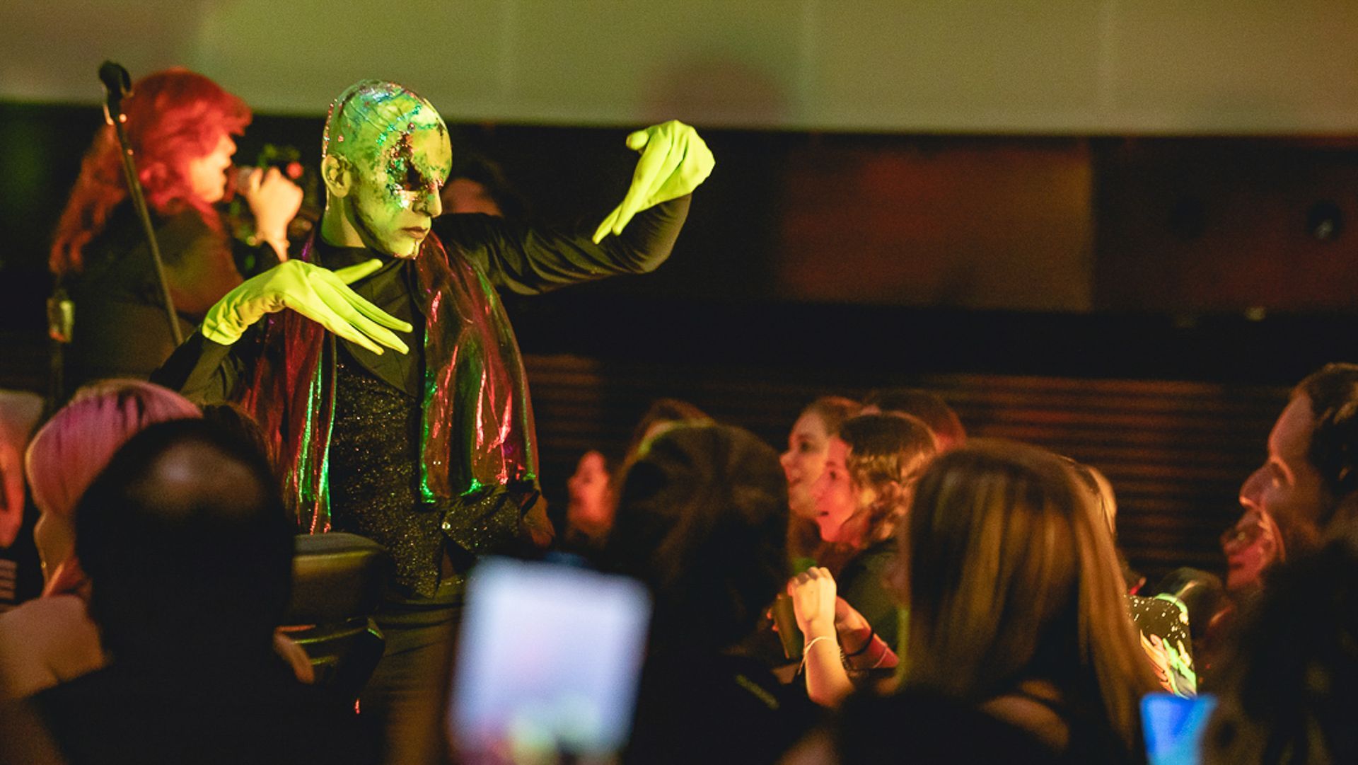 Uma artista com pele verde e luvas brilhantes gesticula expressivamente para o público em um local fechado e com pouca luz.