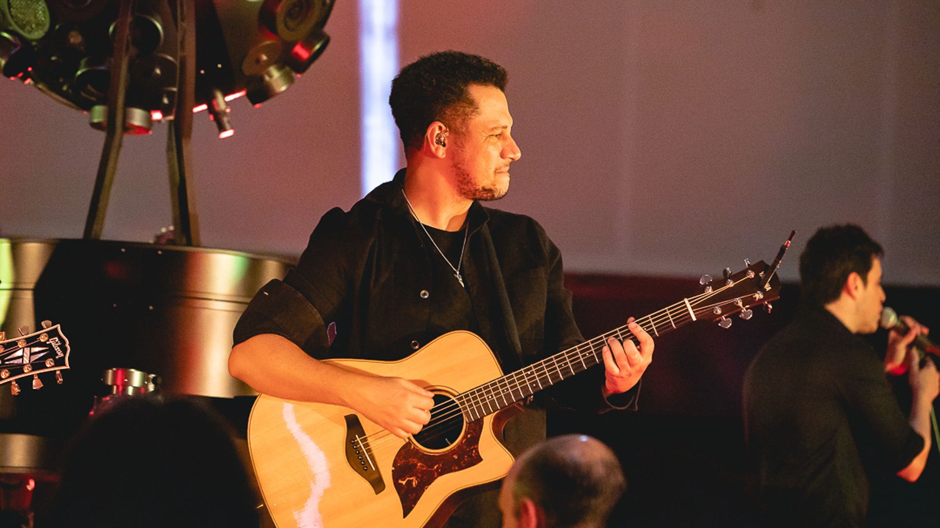 Um artista tocando violão enquanto outra canta ao microfone em um palco com iluminação aconchegante.