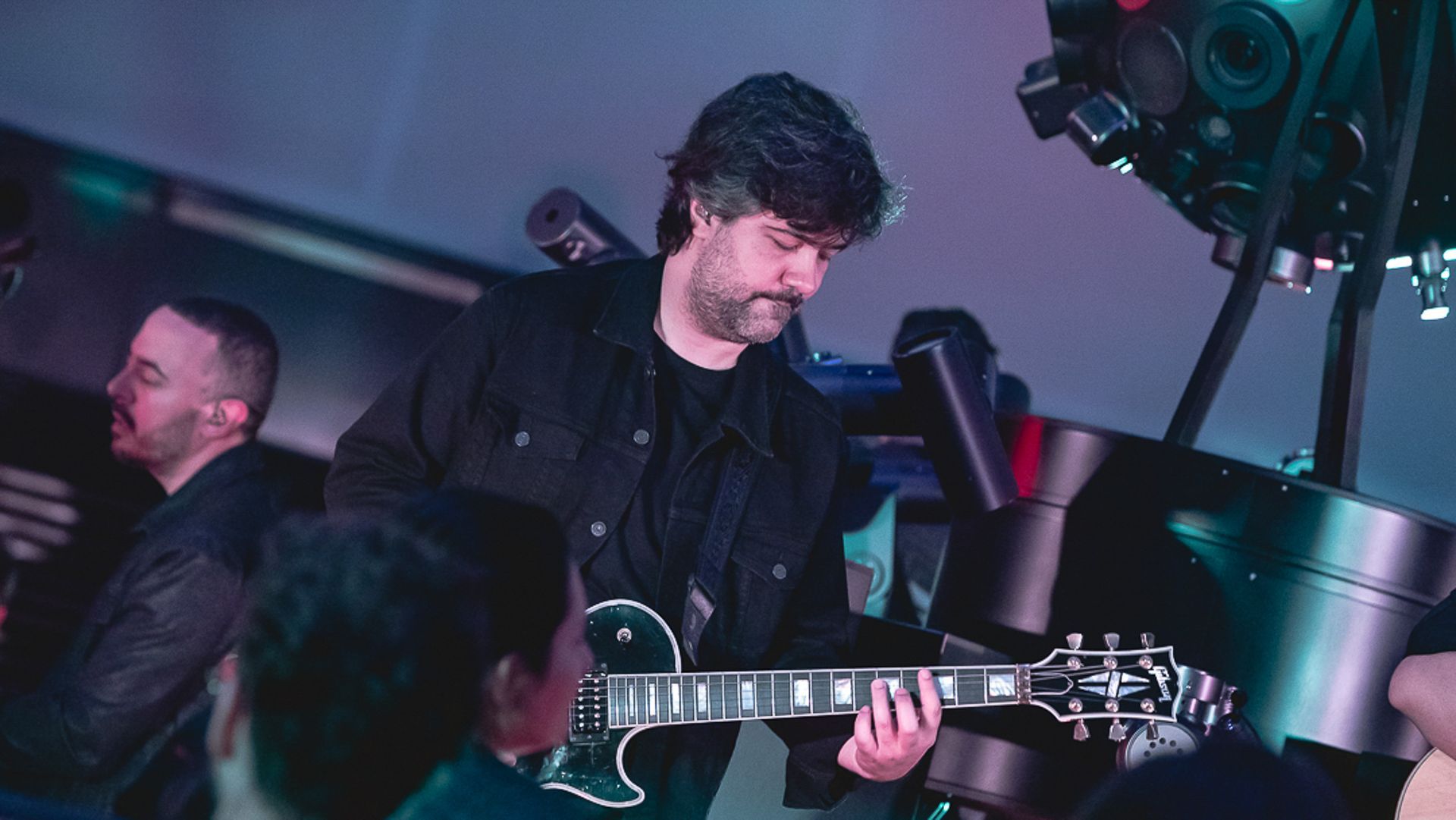 Um músico com jaqueta escura toca uma guitarra elétrica preta enquanto se apresenta no palco sob iluminação cênica.