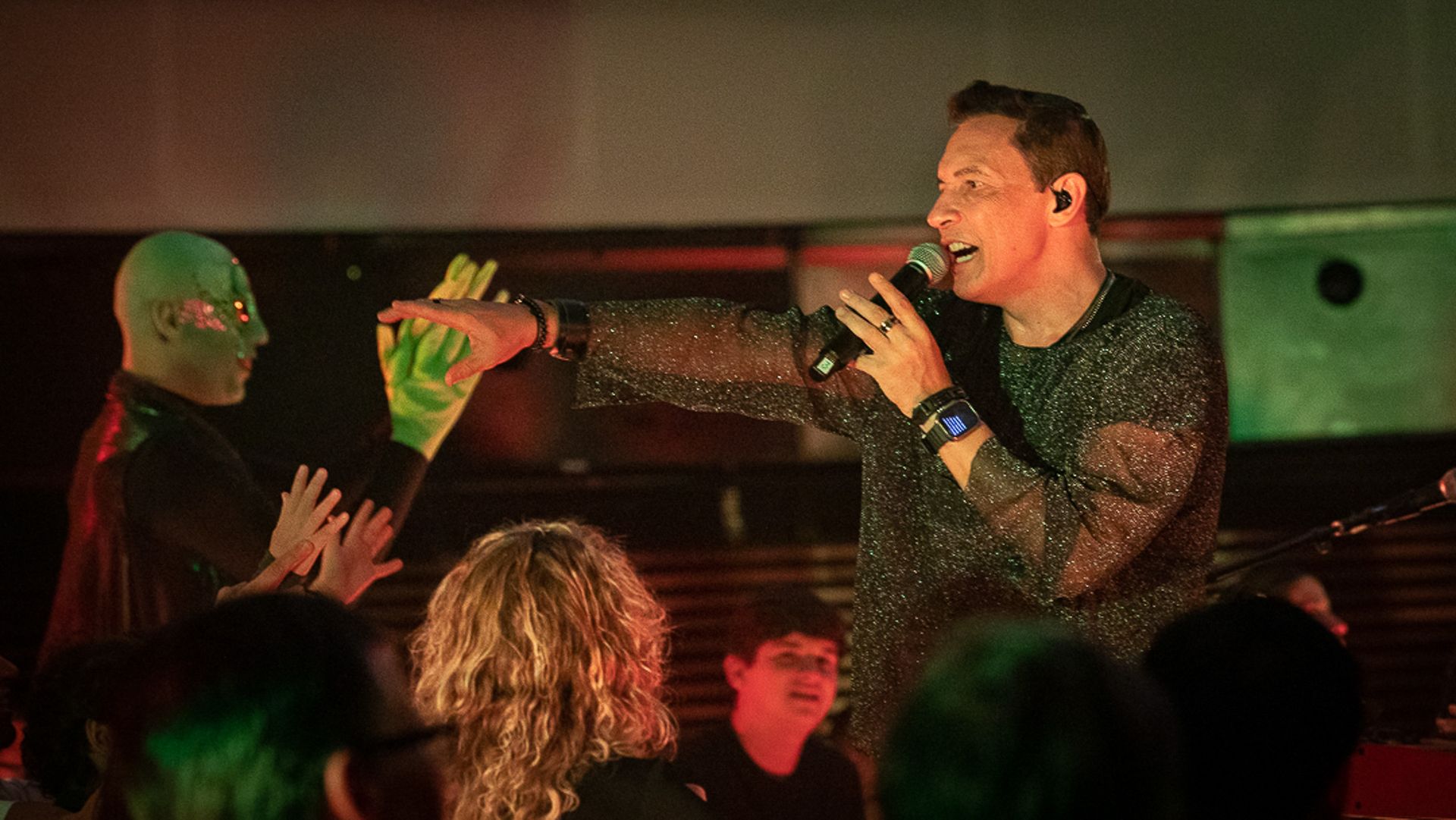 Uma artista que canta ao microfone estende a mão para tocar a mão de uma pessoa com maquiagem verde no rosto e nas mãos.