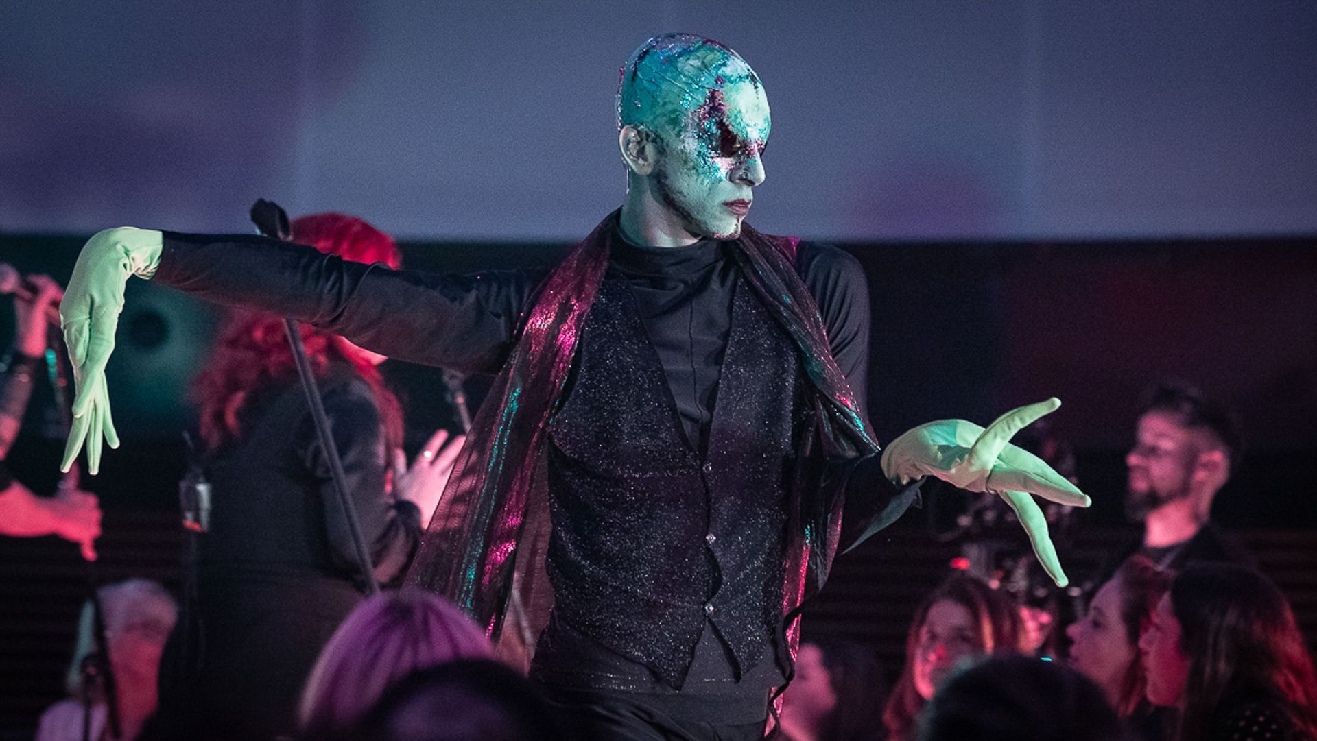 Uma artista com o rosto pintado de verde-azulado, luvas e um colete preto brilhante dança no palco diante de uma plateia.