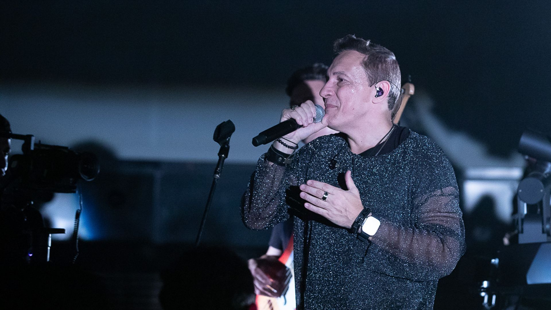 Uma pessoa vestindo uma camisa preta brilhante de mangas compridas canta em um microfone em um palco com um fundo desfocado.