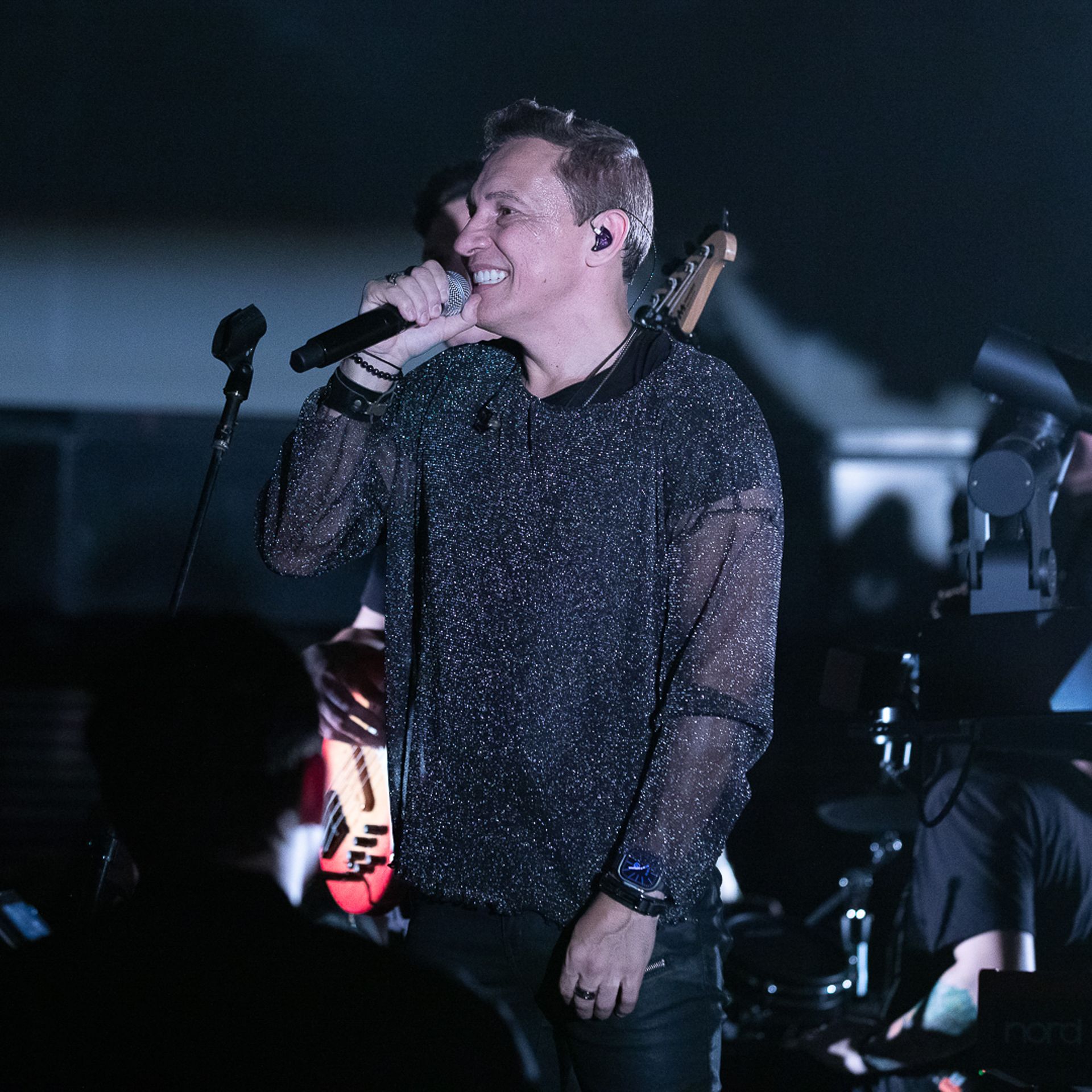 Uma pessoa sorridente cantando em um microfone no palco, vestindo uma blusa preta, semitransparente e brilhante.