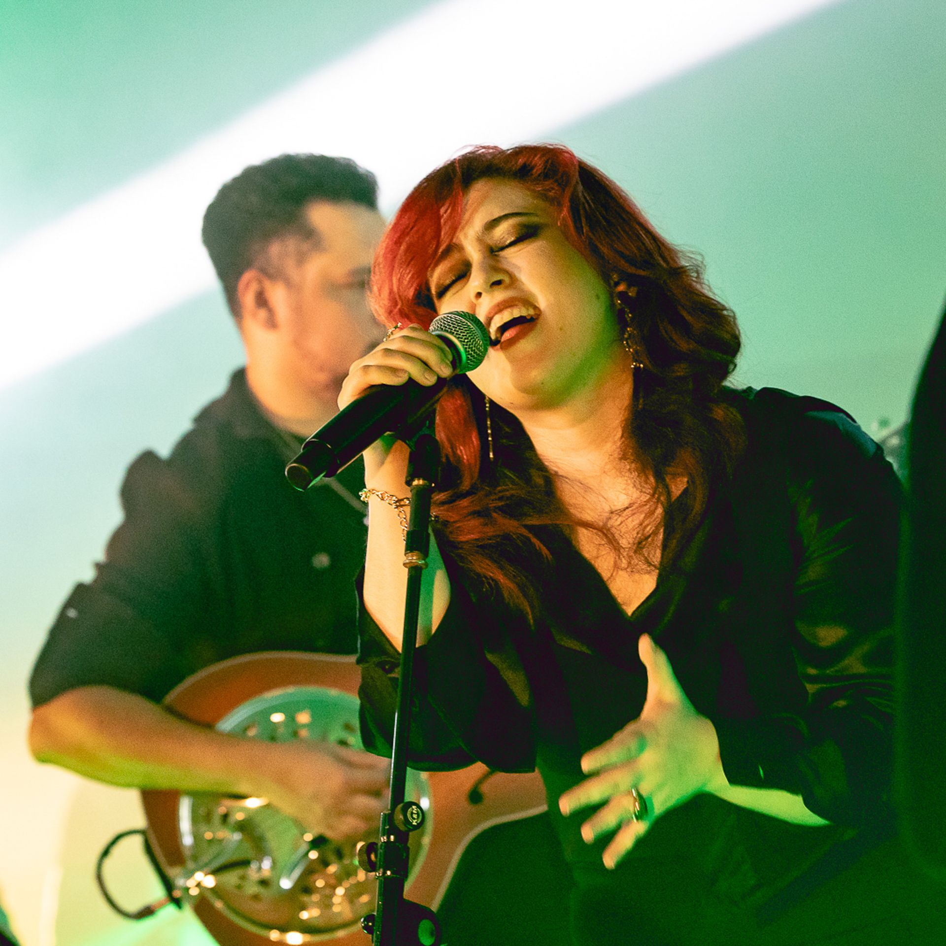 Uma artista canta ao microfone com os olhos fechados, enquanto um guitarrista toca ao fundo, sob luz verde.