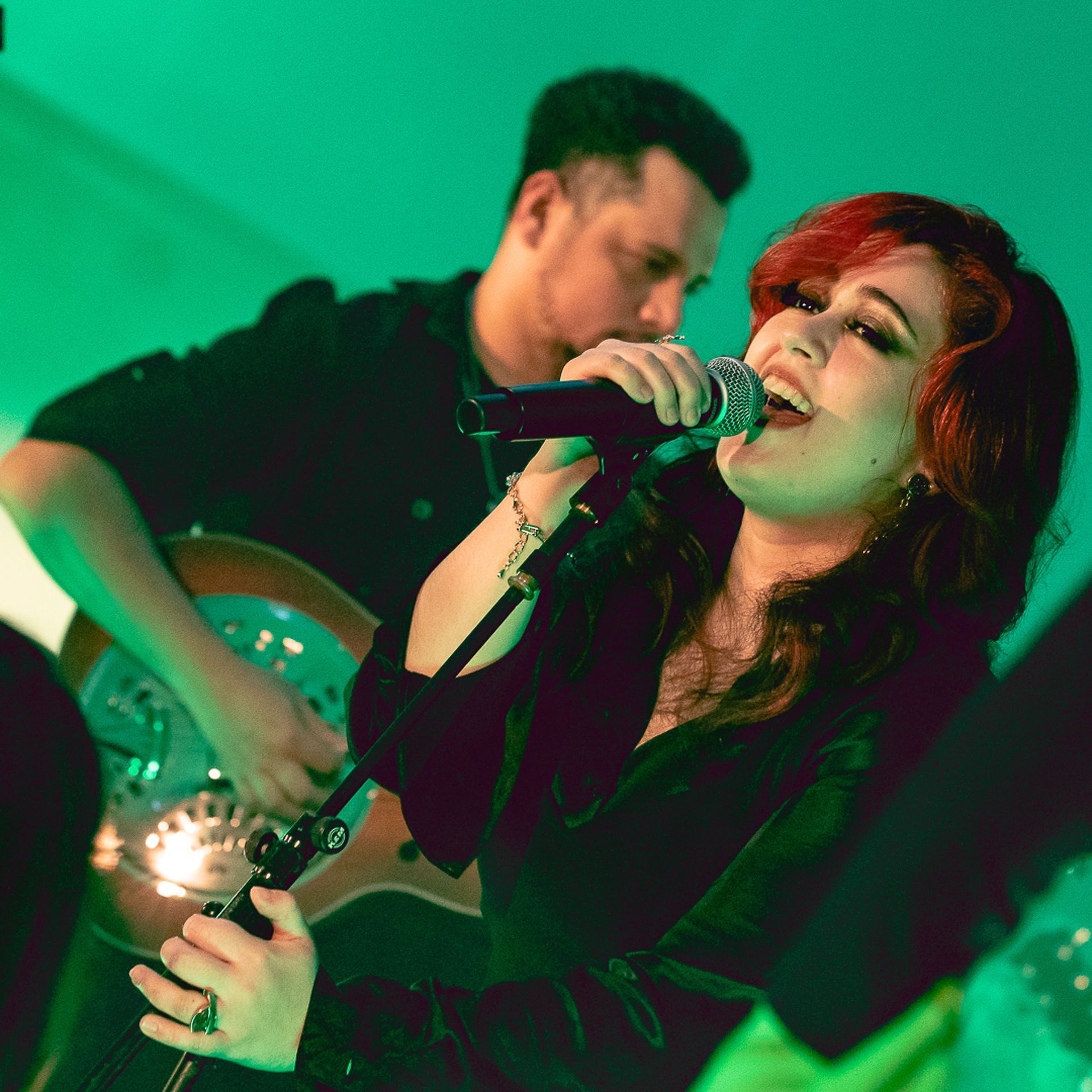 Uma artista canta em um microfone enquanto outra toca um violão ressonador, iluminada por luzes de palco verdes.