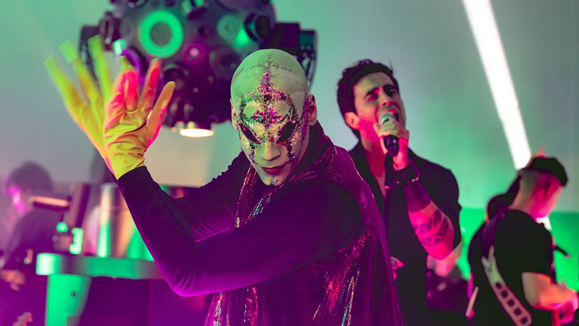 Um artista com máscara branca e luvas amarelas posa em frente a um cantor que segura um microfone sob luzes verdes de palco.