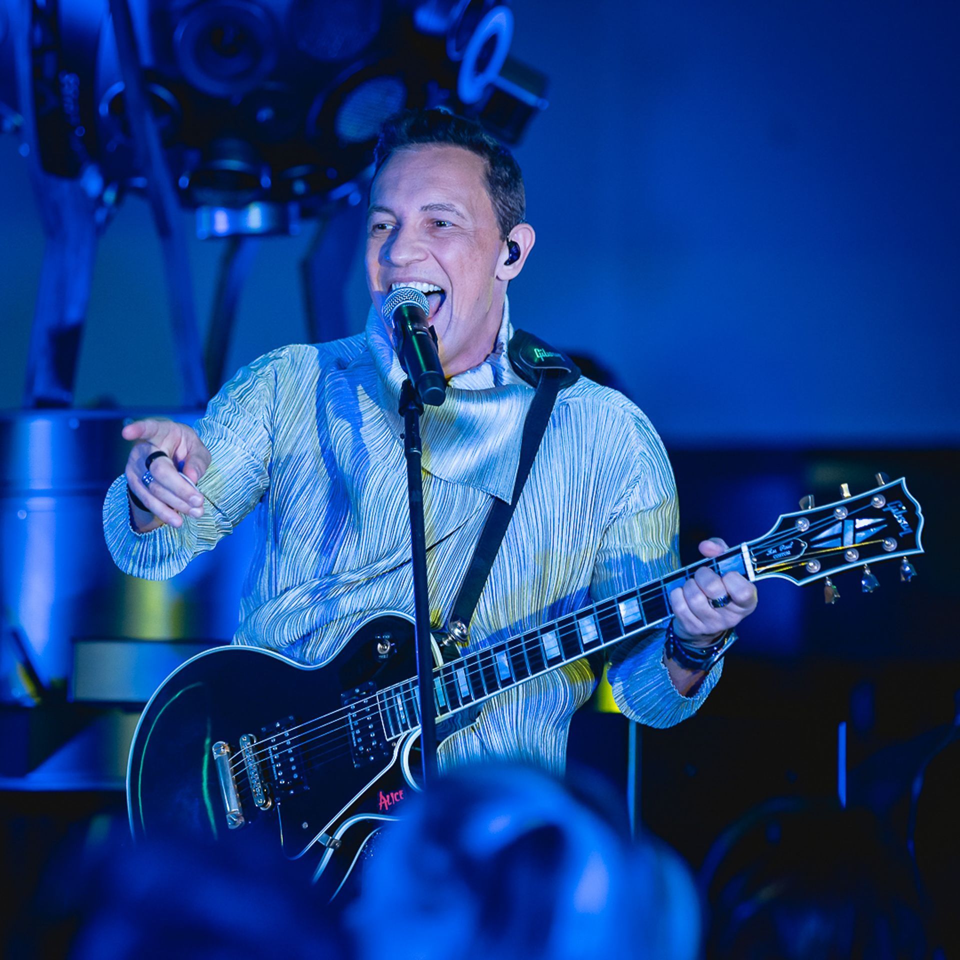 Uma artista sorridente canta ao microfone enquanto toca guitarra elétrica sob luzes azuis de palco.