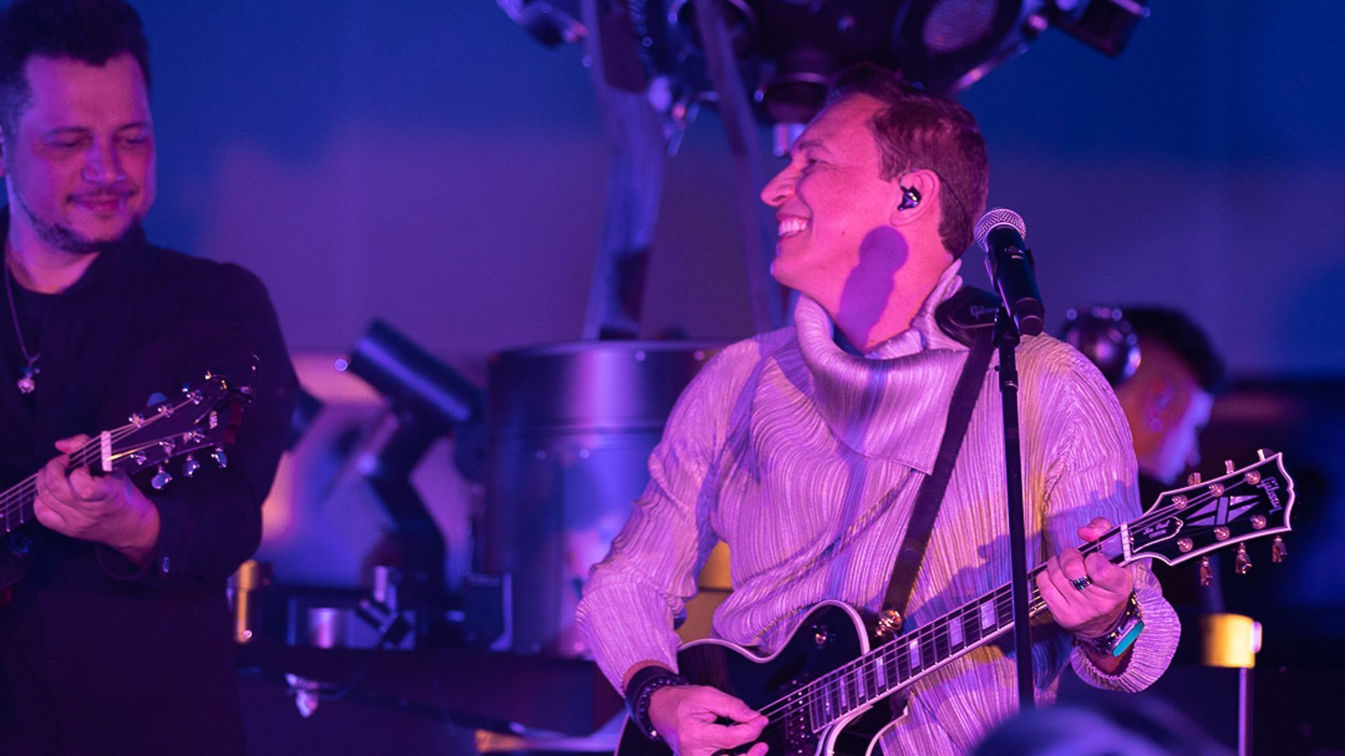 Dois músicos se apresentam no palco sob iluminação azul e roxa; um toca violão enquanto o outro sorri.