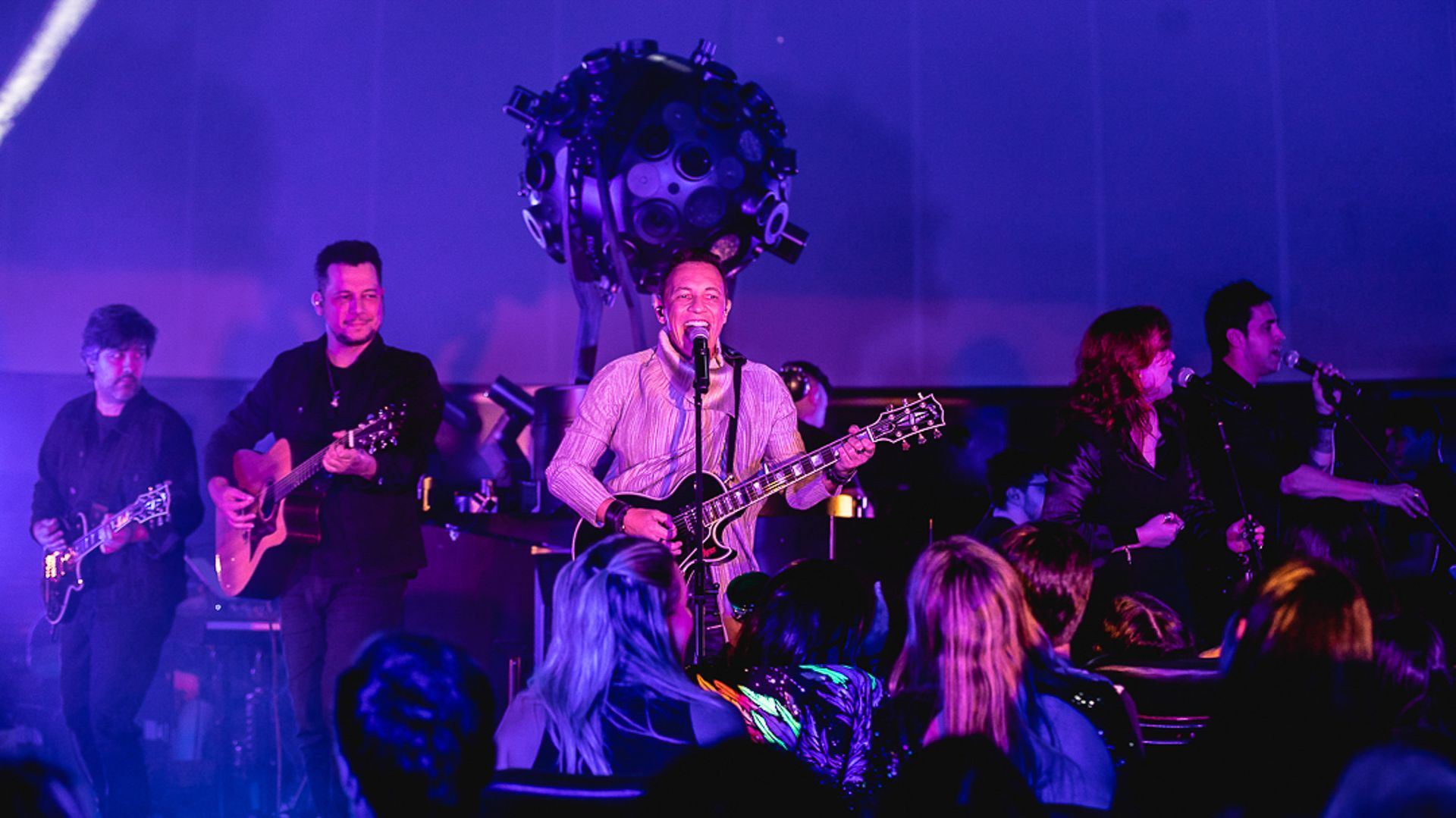 Uma banda se apresenta no palco sob luzes roxas, com um vocalista tocando guitarra e cantando para a plateia.