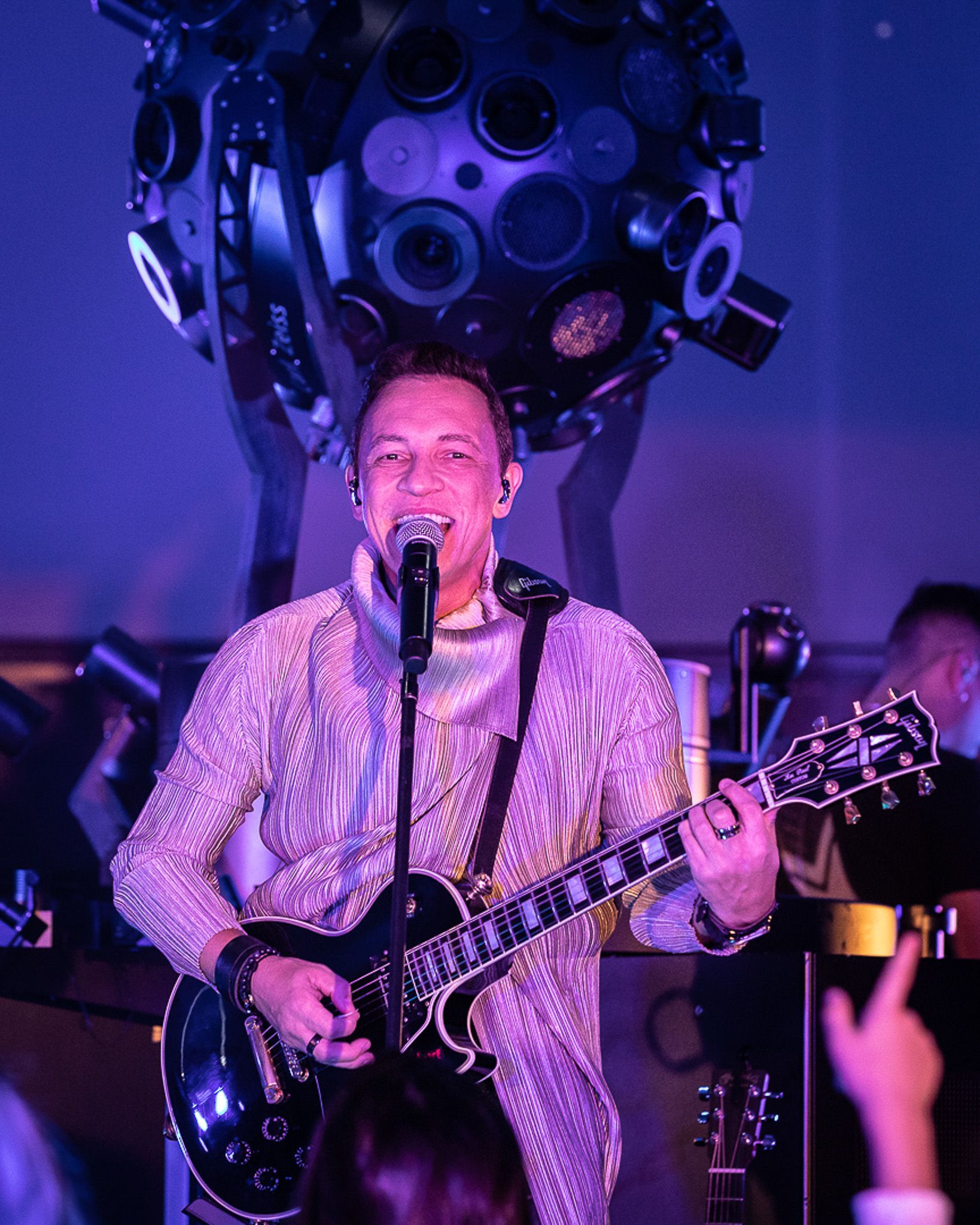 Um artista canta ao microfone enquanto toca guitarra elétrica sob luzes de palco roxas.