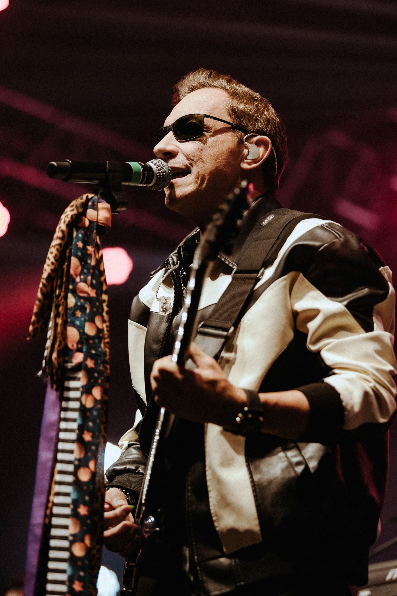 Um artista de óculos escuros tocando violão e cantando ao microfone no palco. Ele veste uma jaqueta preta e branca.