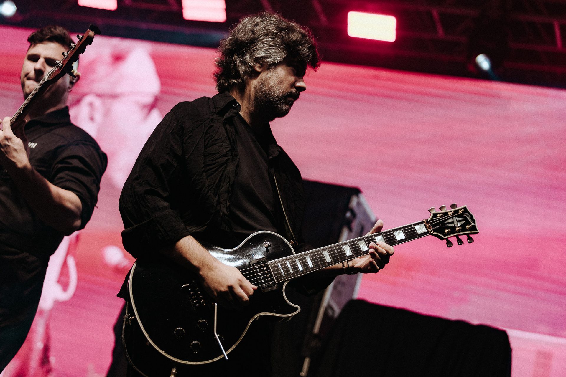 Guitarristas se apresentando no palco, um deles vestido de preto com cabelos grisalhos, tocando uma guitarra preta.
