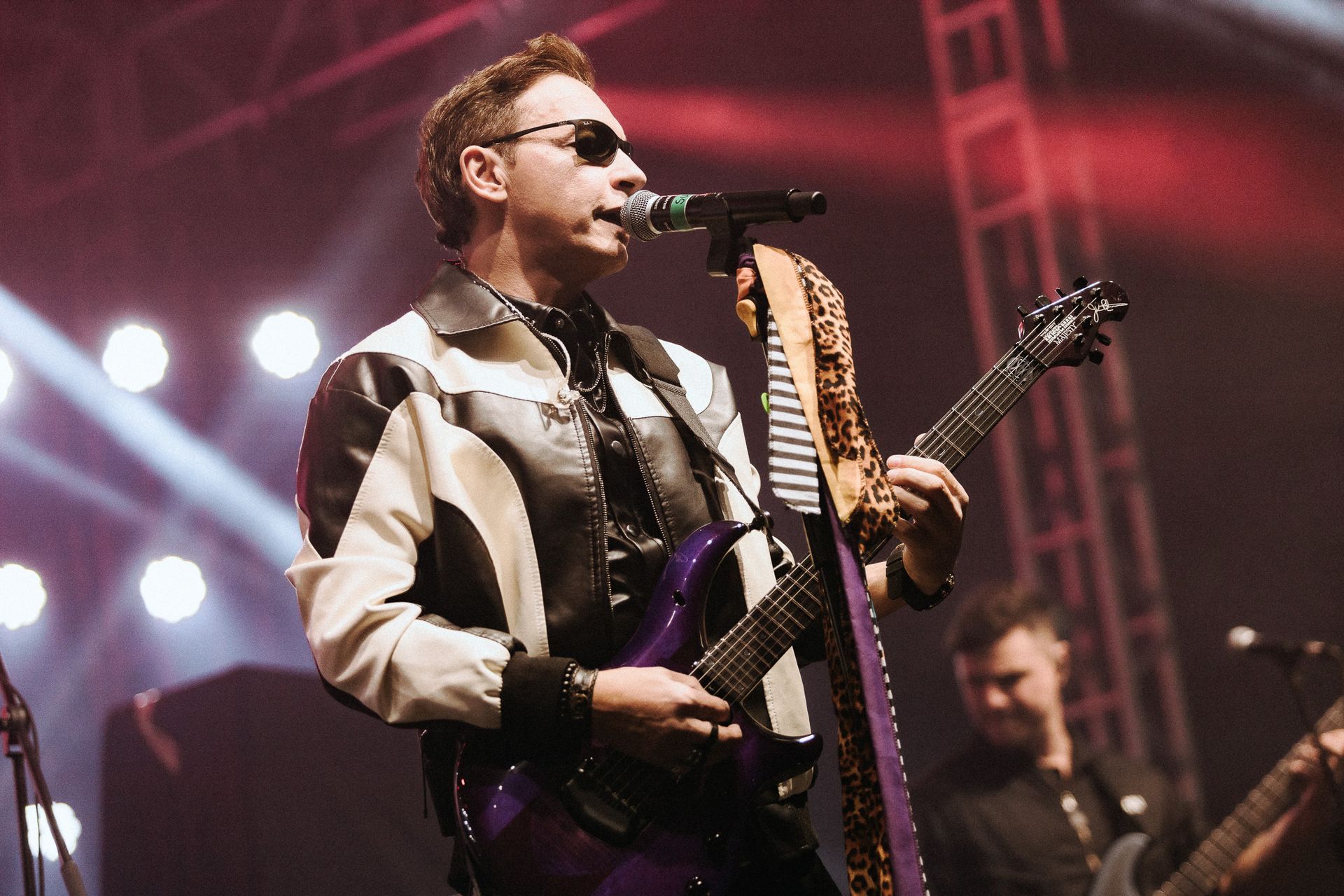 Um artista de óculos escuros tocando guitarra roxa no palco, cantando ao microfone.