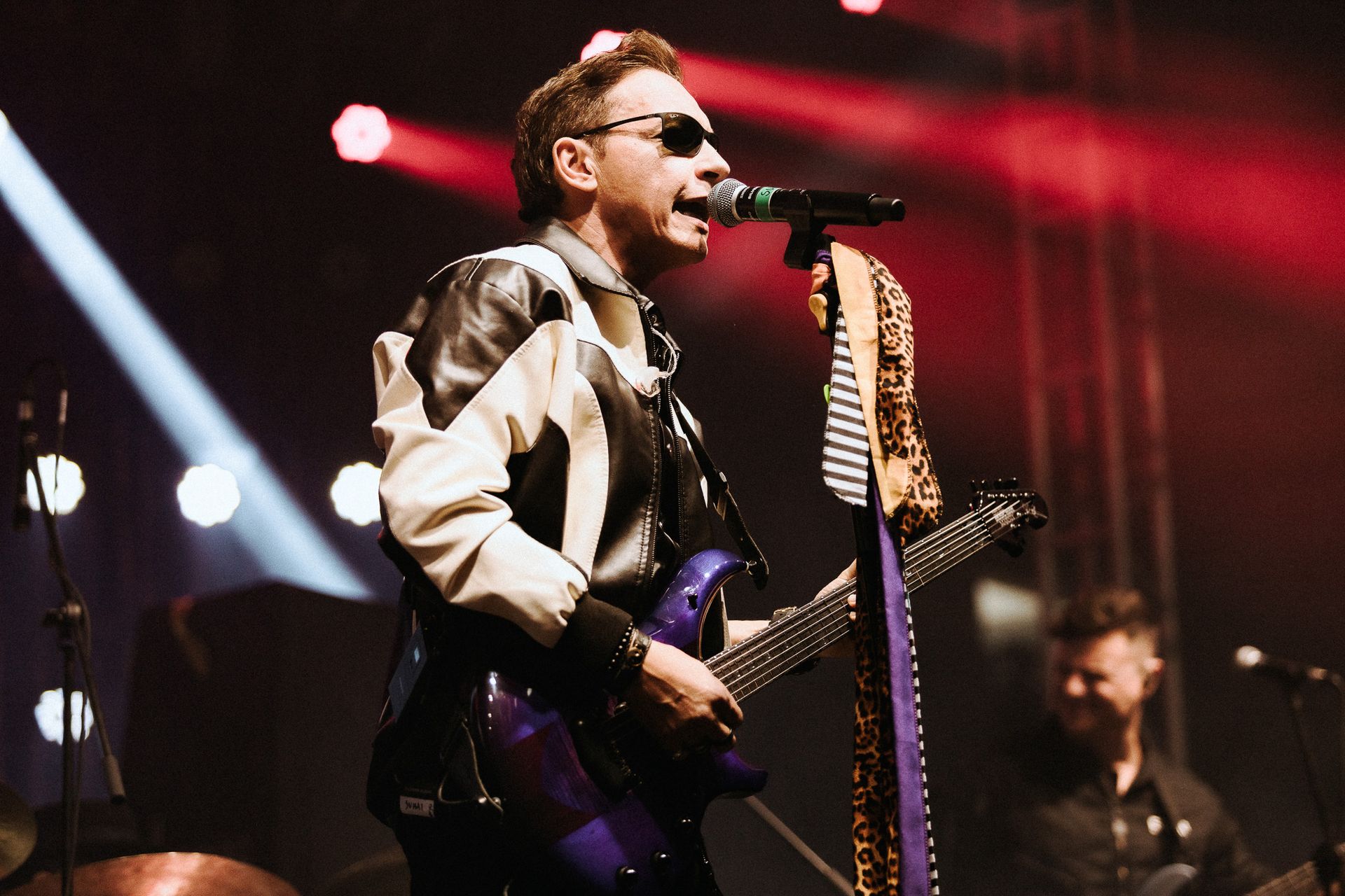 Um artista cantando e tocando violão no palco, usando óculos escuros e uma jaqueta preta e branca.