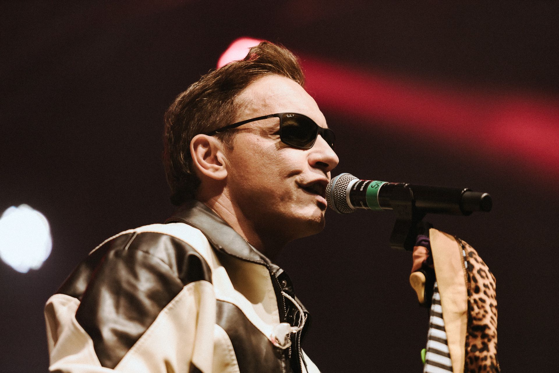 Cantor de óculos escuros se apresentando com um microfone, vestindo jaqueta, com luzes de palco ao fundo.