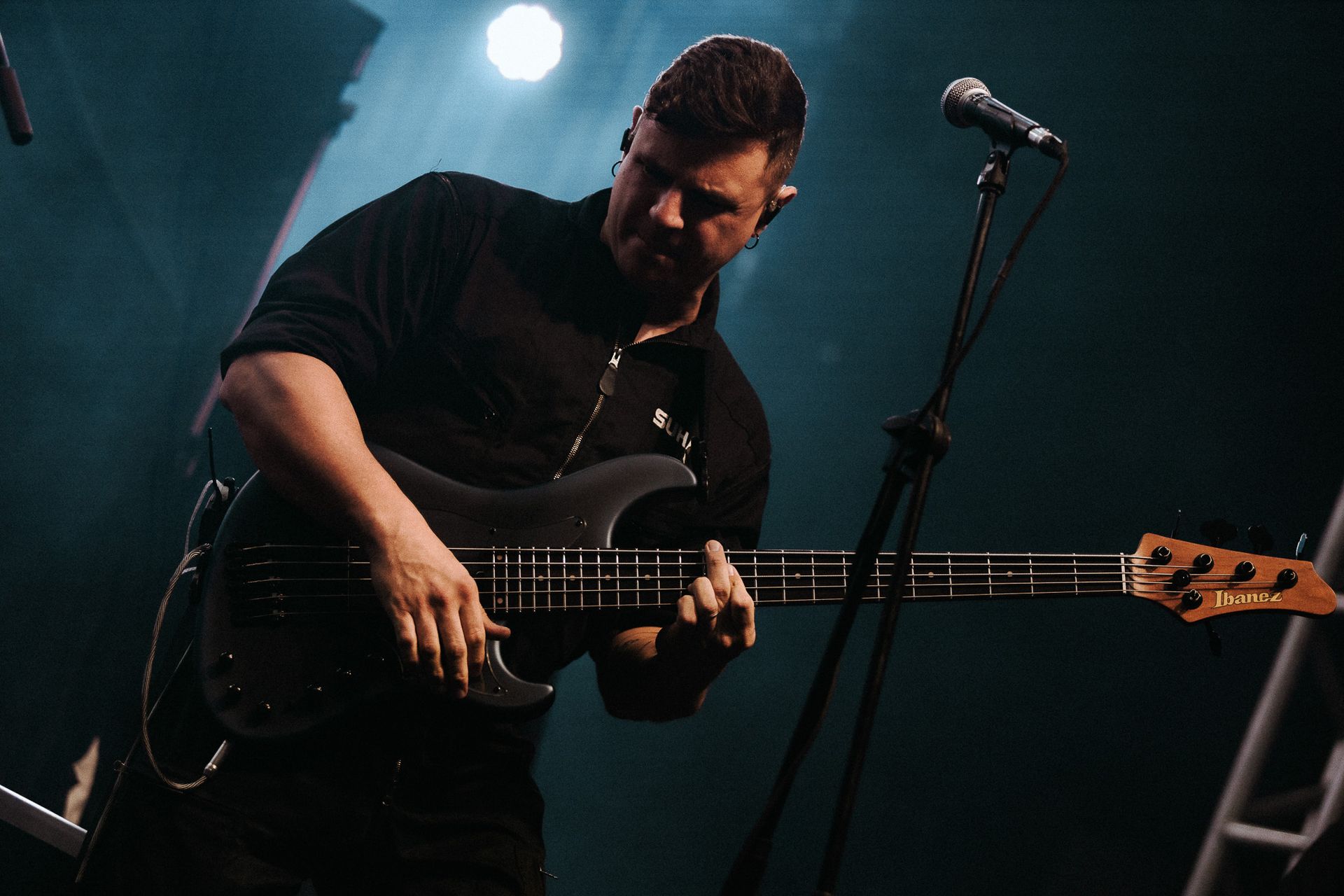 Um artista tocando um baixo preto no palco com um microfone. Ambiente escuro com iluminação de palco.