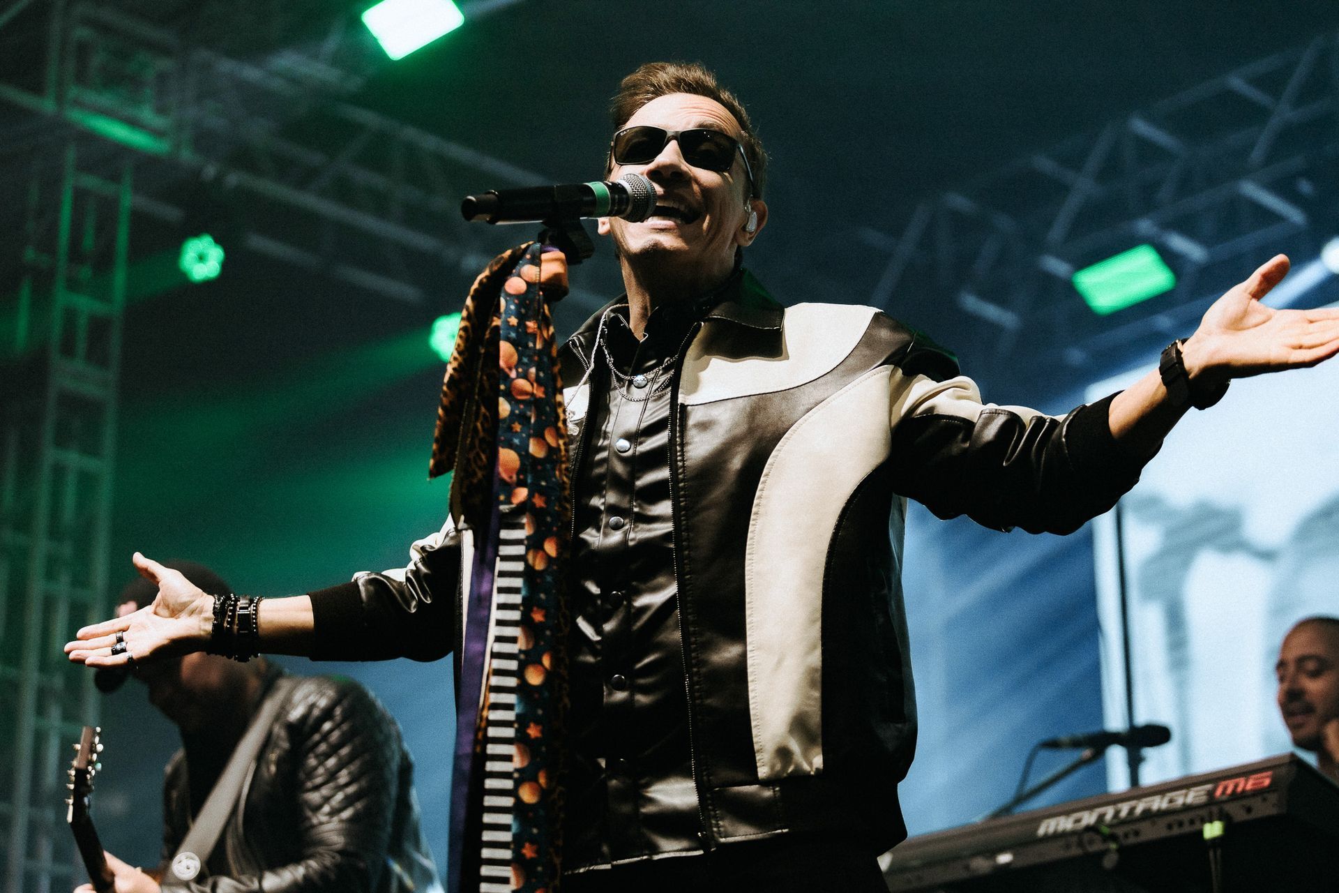 Cantor no palco com os braços estendidos, usando óculos escuros e jaqueta preta e branca. Iluminação de palco verde.