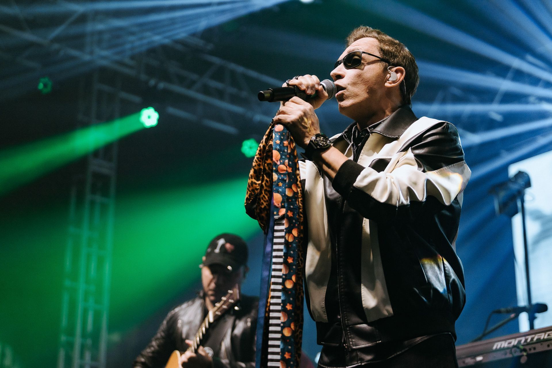 Cantor se apresentando no palco, segurando um microfone, usando óculos escuros e uma jaqueta preta e branca.