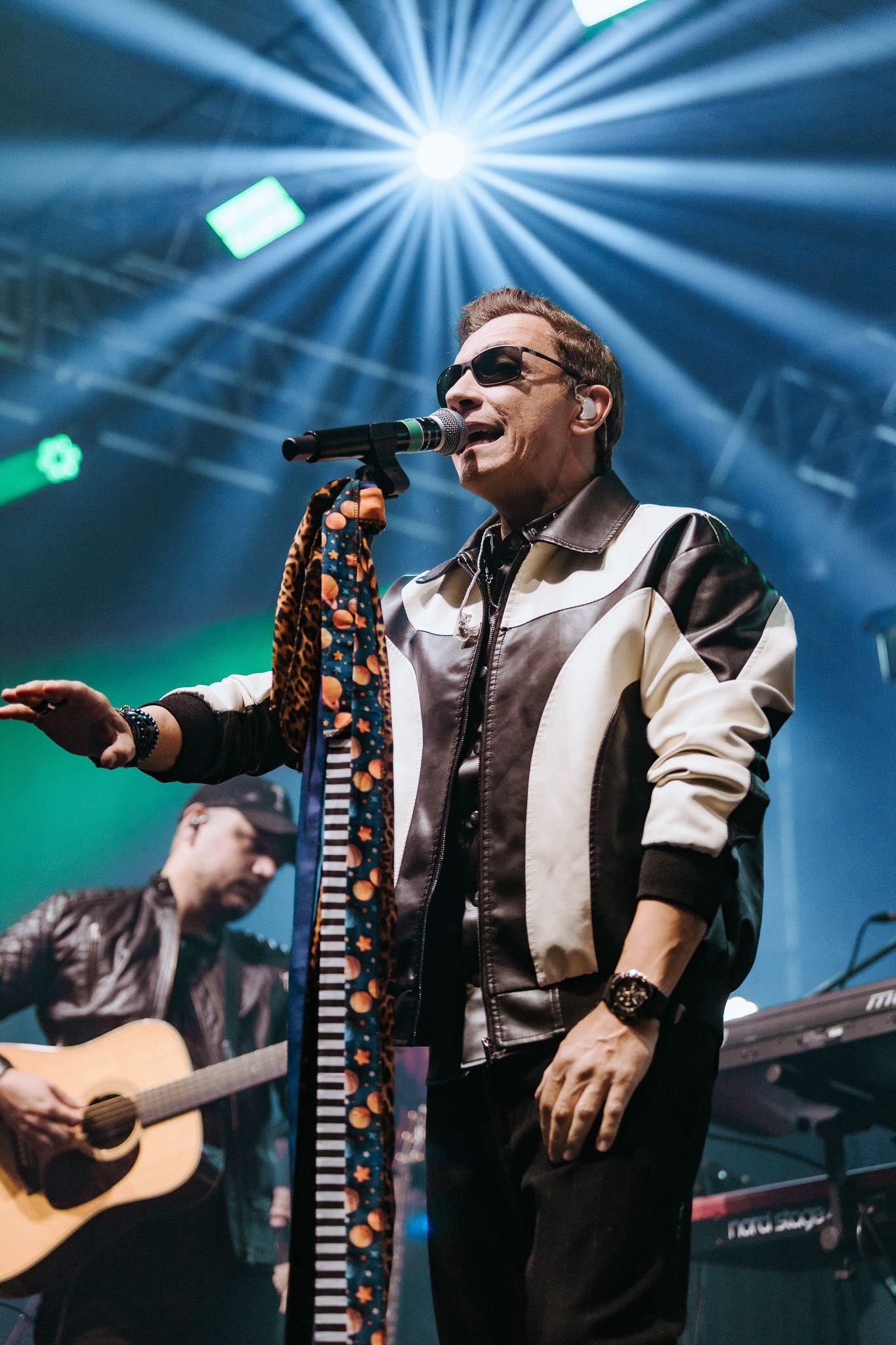 Um artista cantando ao microfone no palco, vestindo uma jaqueta preta e branca. Guitarrista ao fundo.